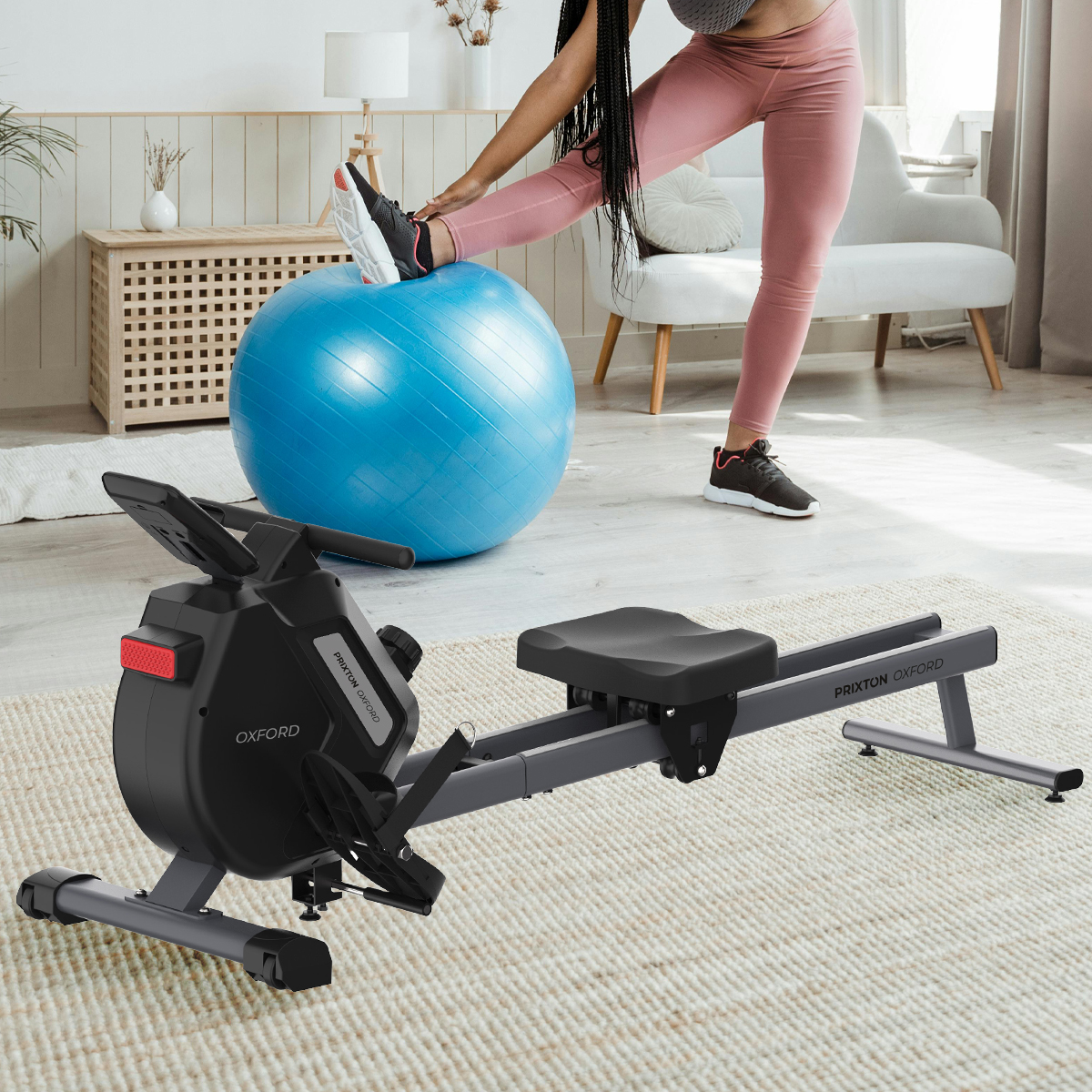 Kobieta rozciąga się na wioślarzu. Niebieska piłka i biała sofa w tle. Pokój z dywanem.