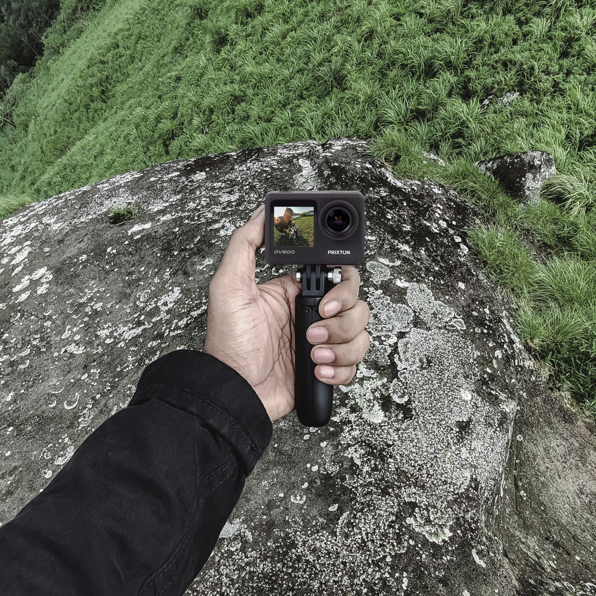 Ręka trzymająca czarną kamerę sportową na skale, tło z zieloną trawą.