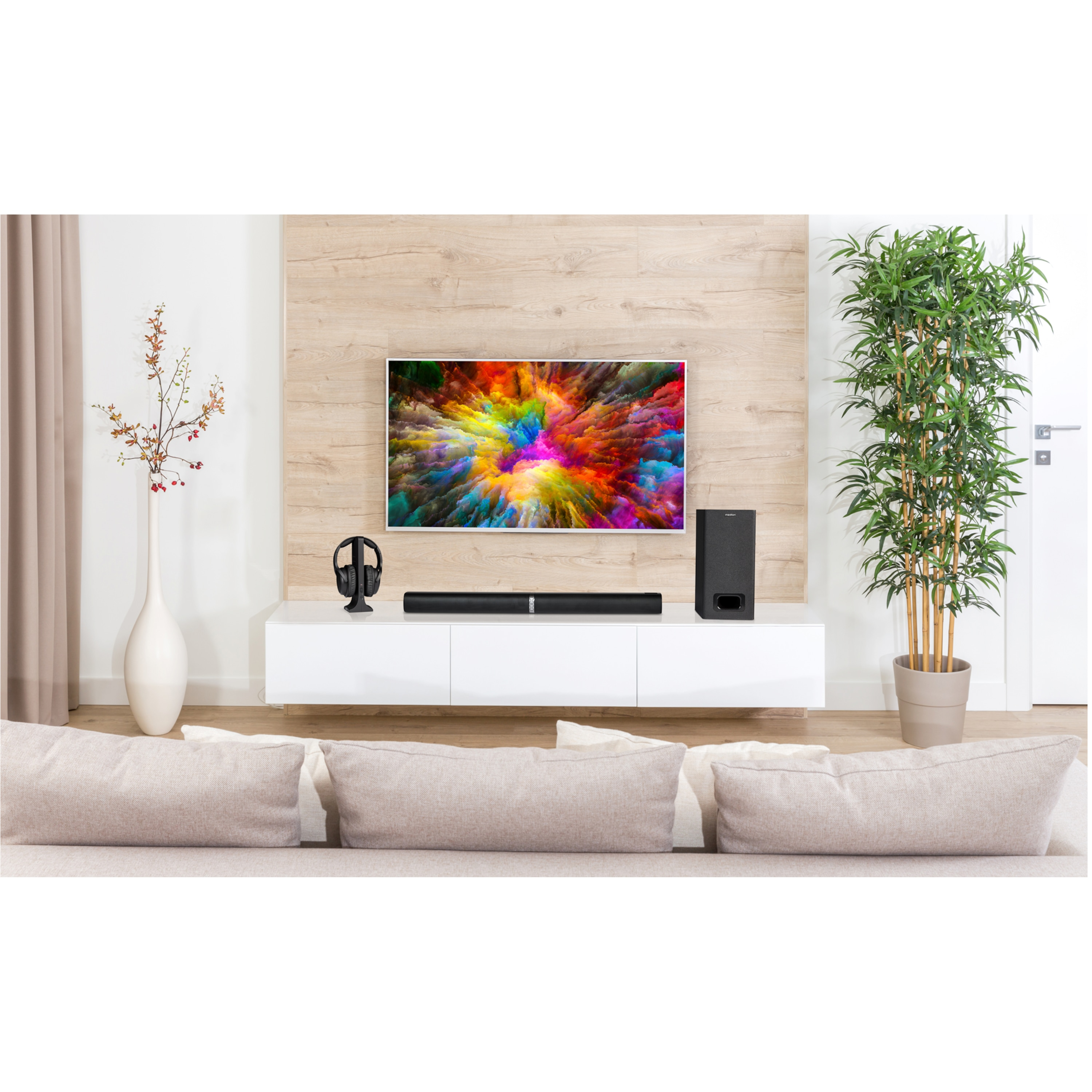 Wohnzimmerszene mit Fernseher, Soundbar und Subwoofer. Couch, Pflanze und Dekorationen sind vorhanden.