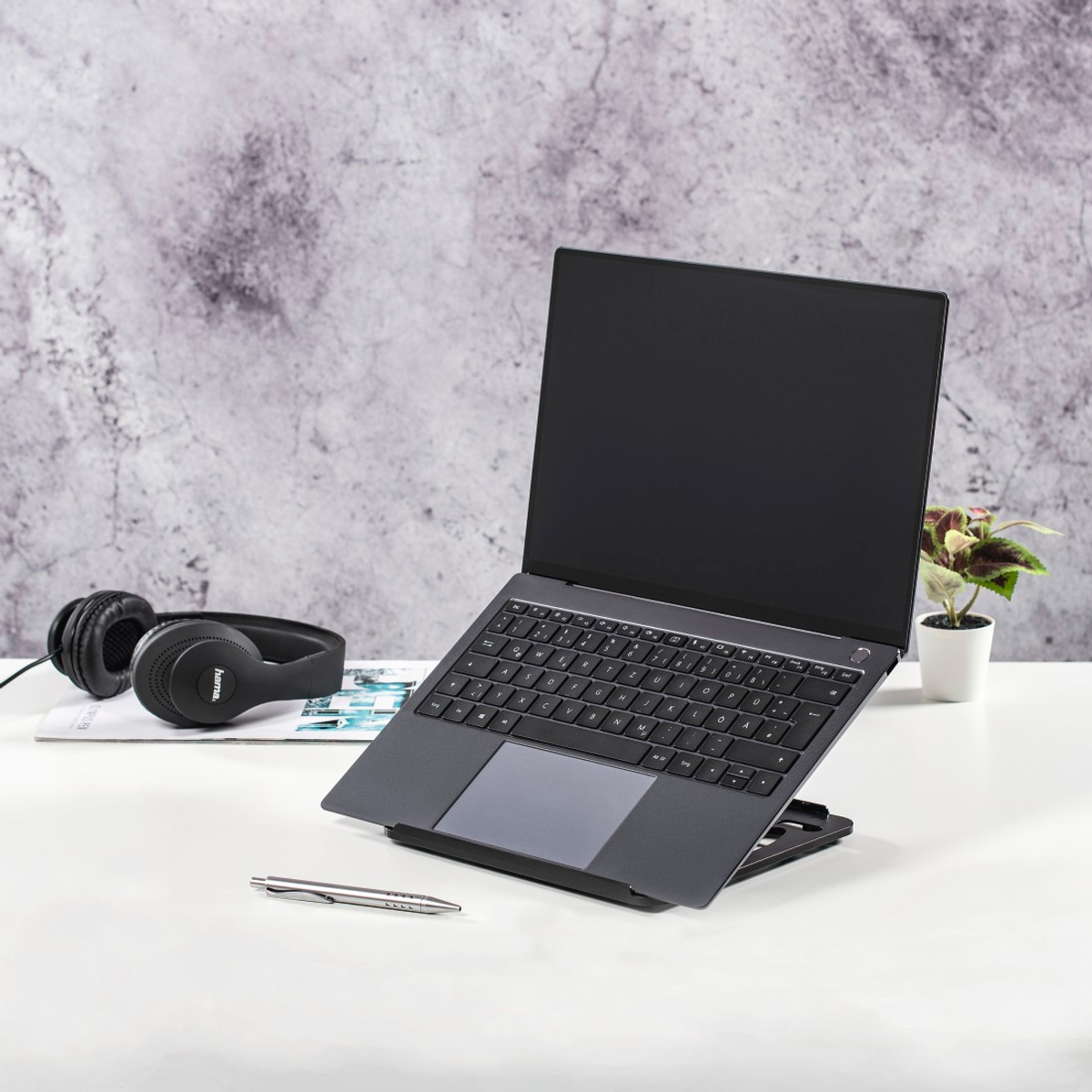 Ein Laptop auf einem Ständer auf einem weißen Schreibtisch. Kopfhörer und ein Stift sind ebenfalls vorhanden. Der Hintergrund ist eine marmorierte graue Wand.