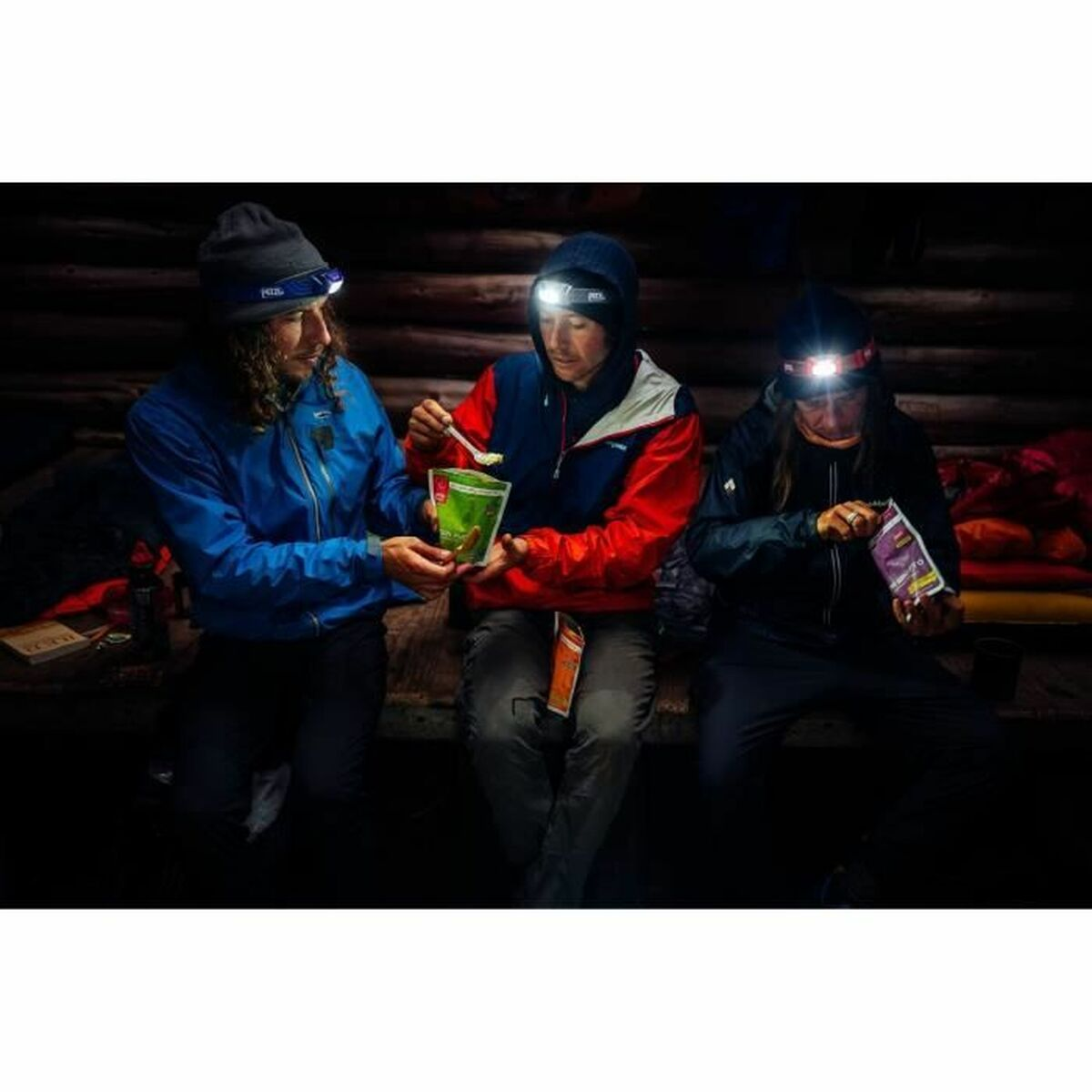 Trzy osoby jedzące z czołówkami w słabo oświetlonej drewnianej chatce.