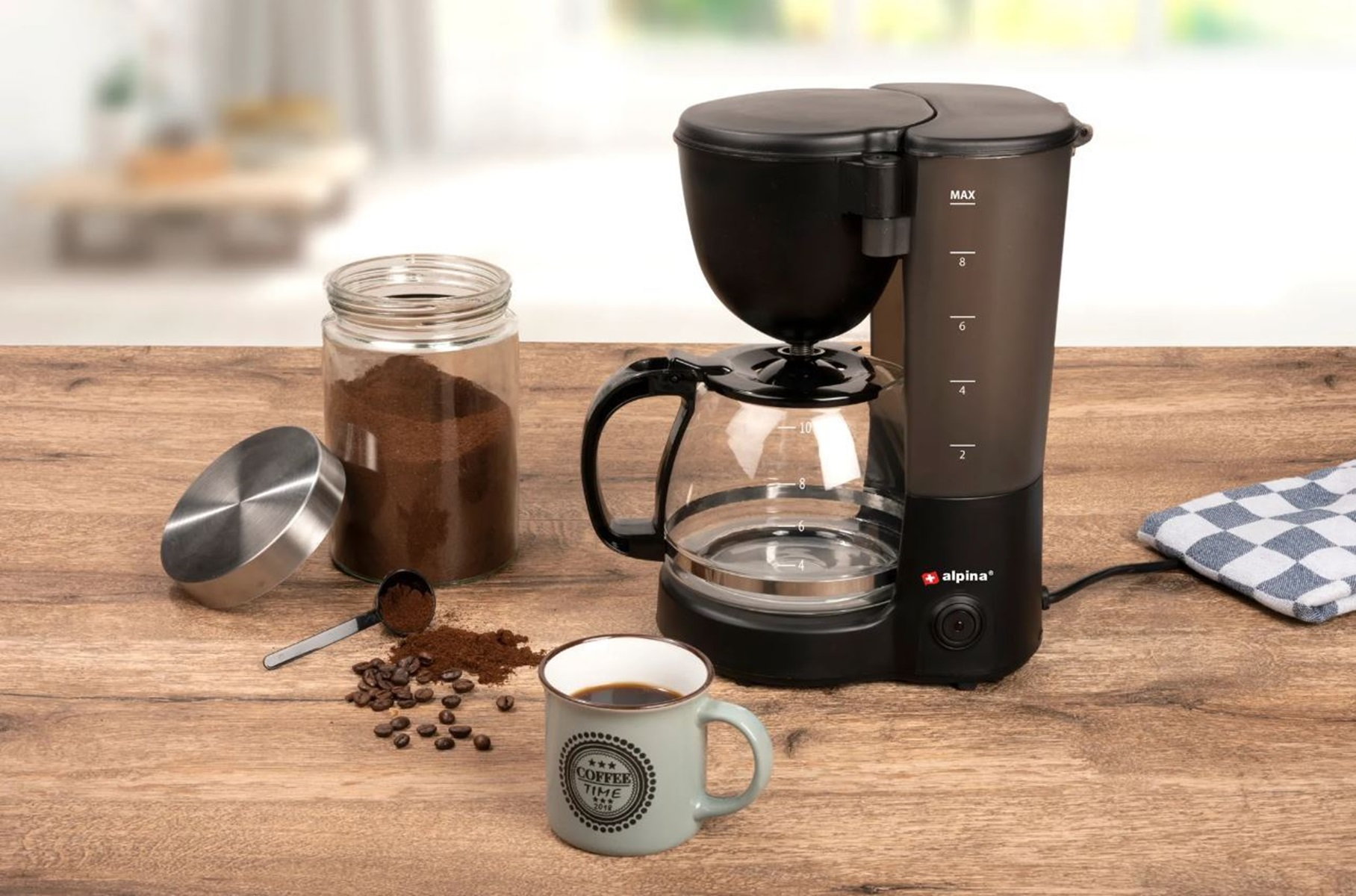 Eine Kaffeemaschine mit Glaskaraffe, Kaffeebohnen, gemahlenem Kaffee, einer Tasse und einem Glas auf einem Holztisch.