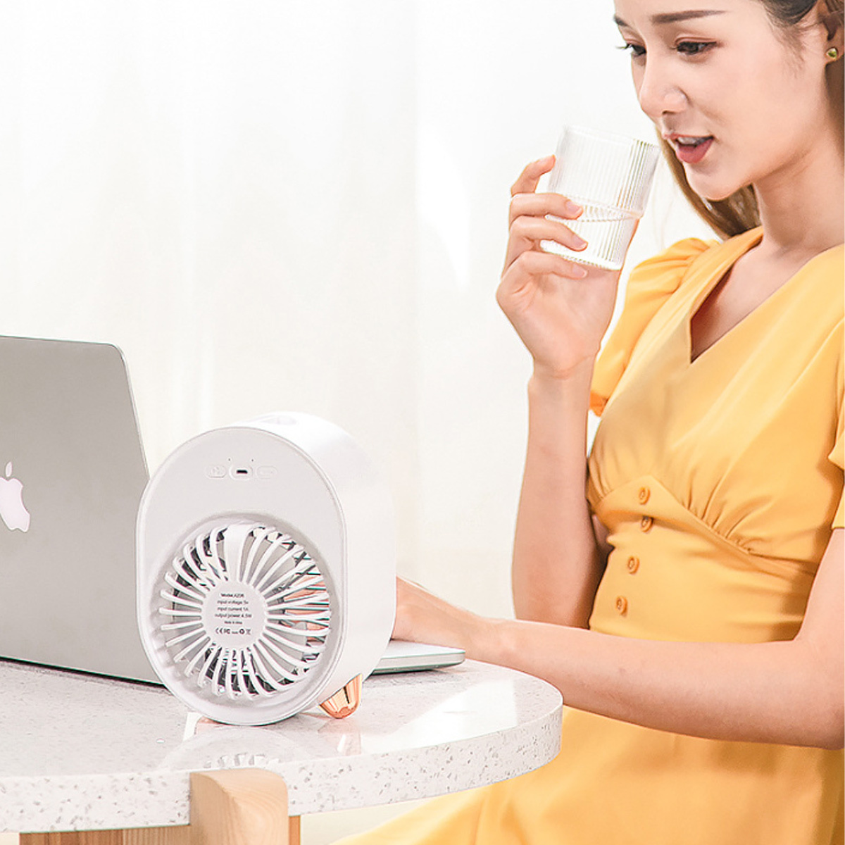 Eine Frau in Gelb trinkt Wasser in der Nähe eines weißen Ventilators und eines Laptops auf einem Holztisch.