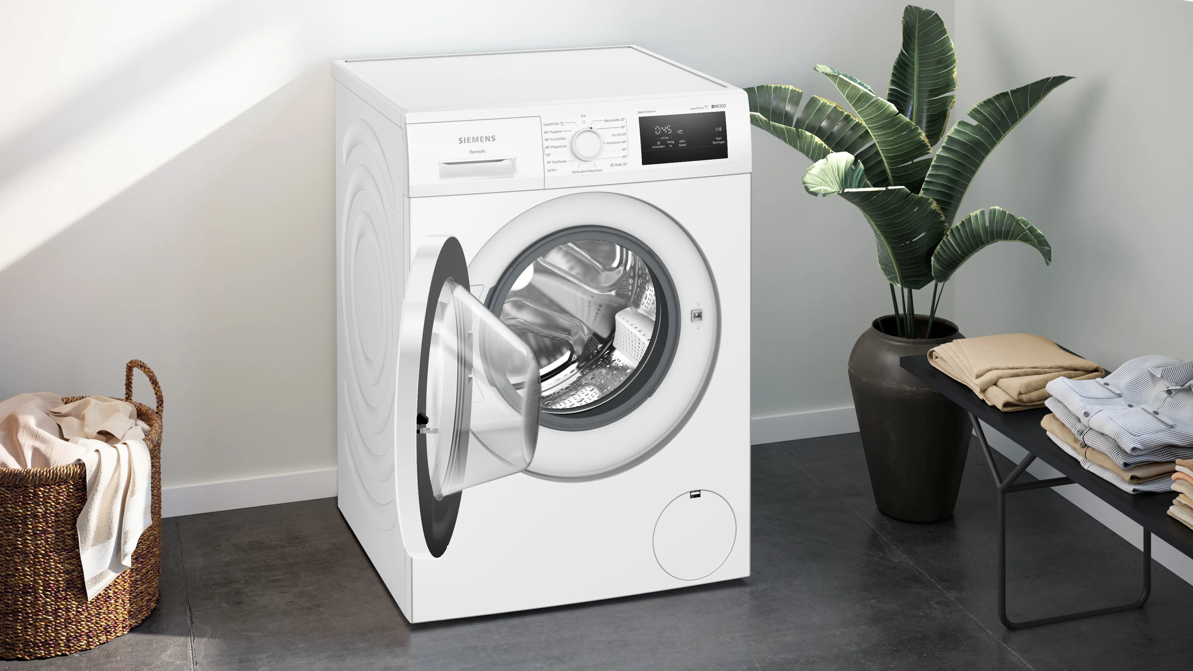 Eine weiße Siemens Waschmaschine mit offener Tür. Ein Korb, Pflanzen und ein Tisch mit Kleidung sind in der Nähe.
