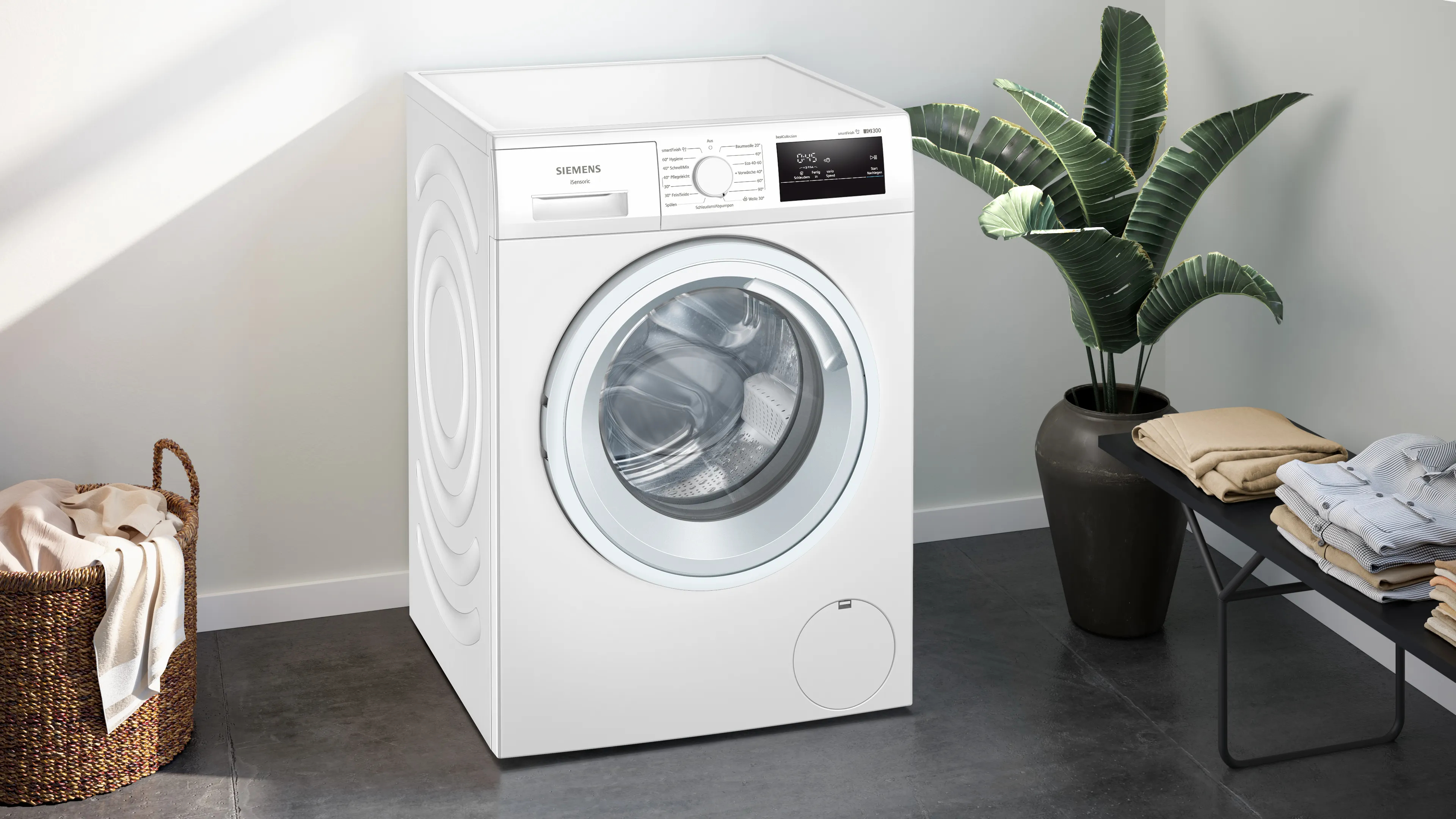 Eine weiße Siemens Waschmaschine in einem Raum mit Pflanze und Bank mit gefalteter Kleidung.