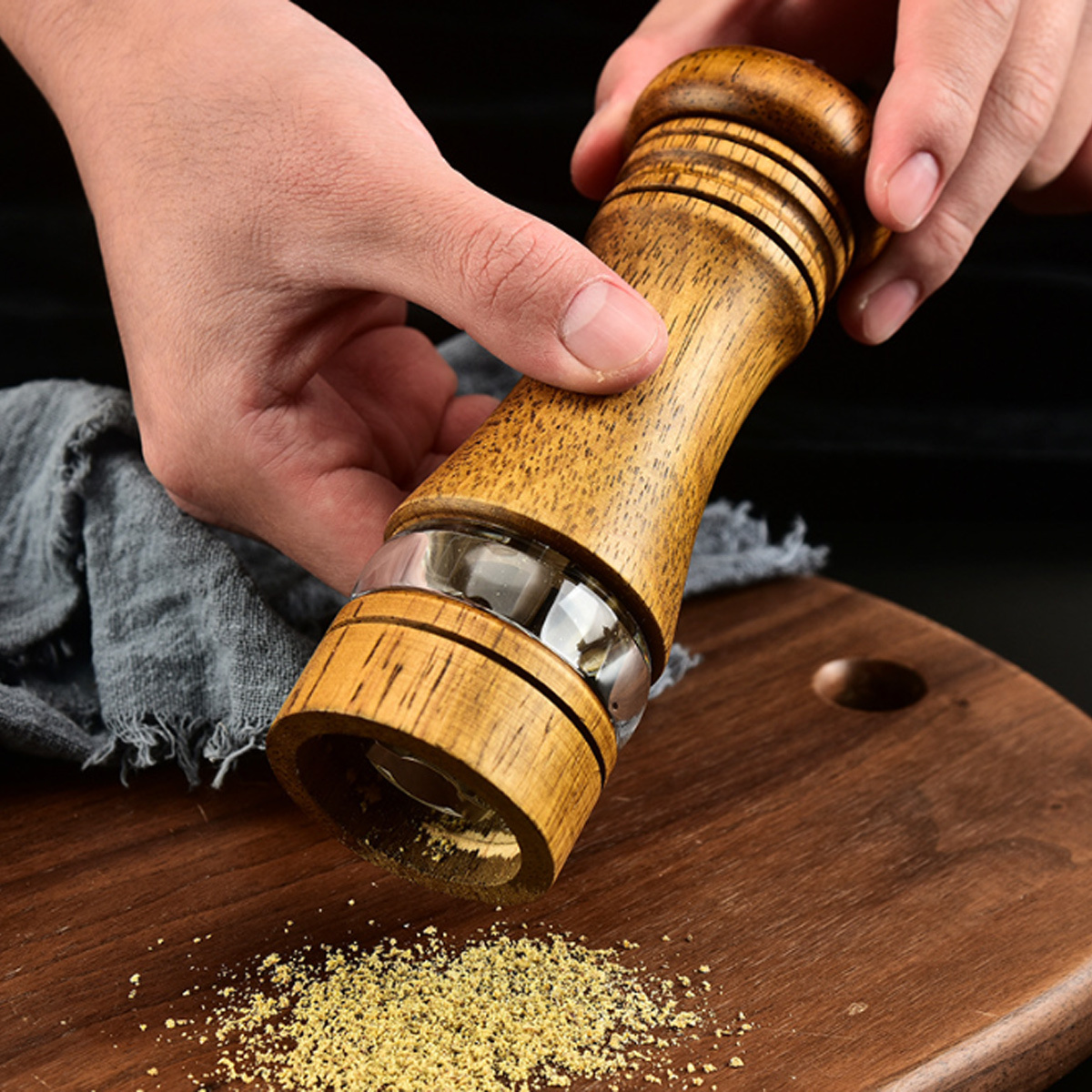 Hände, die Pfeffer mit einer Holzmühle auf einem Holzbrett mit Pfefferpartikeln mahlen.