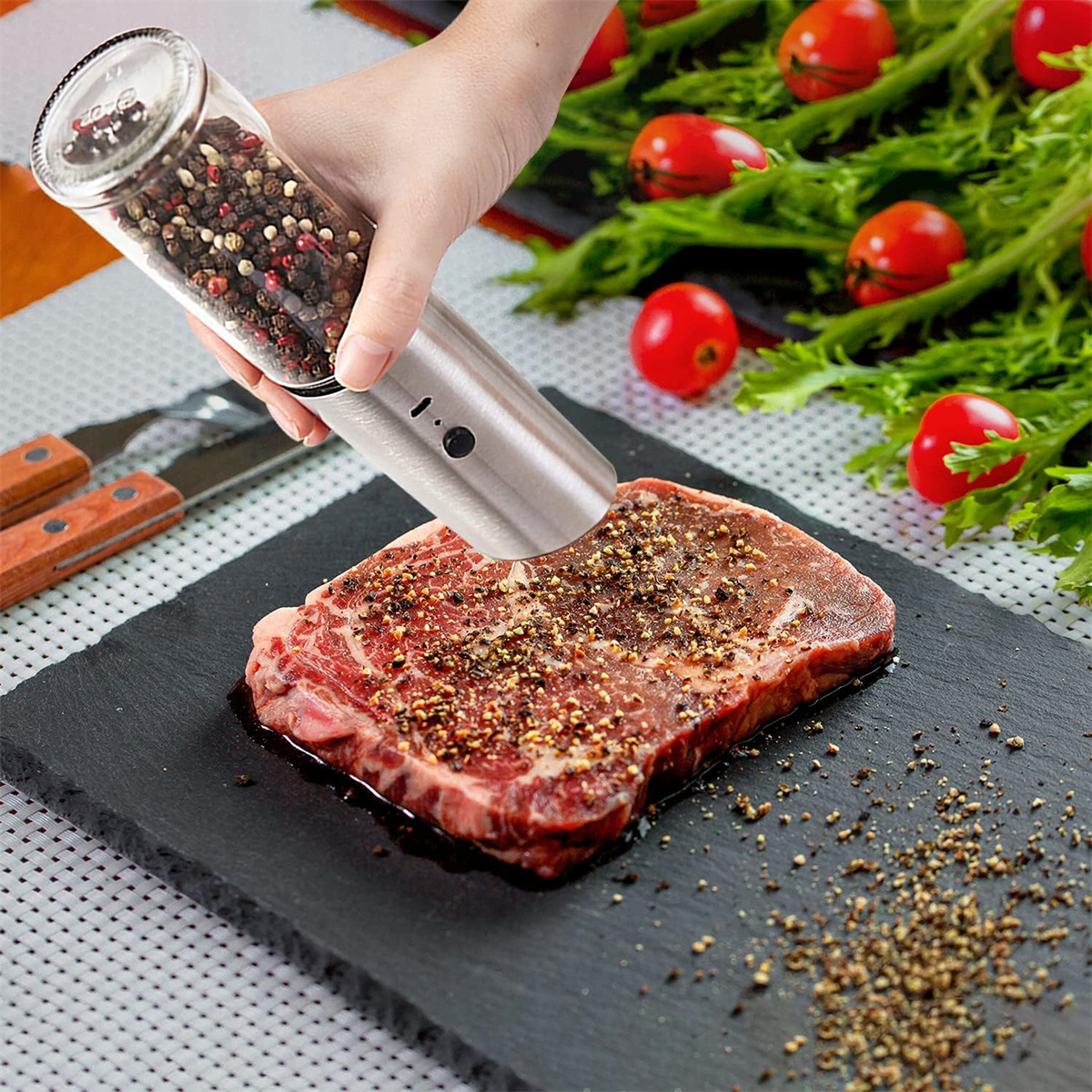 Hand benutzt eine Pfeffermühle auf einem Steak, auf schwarzem Untergrund, Tomaten im Hintergrund.
