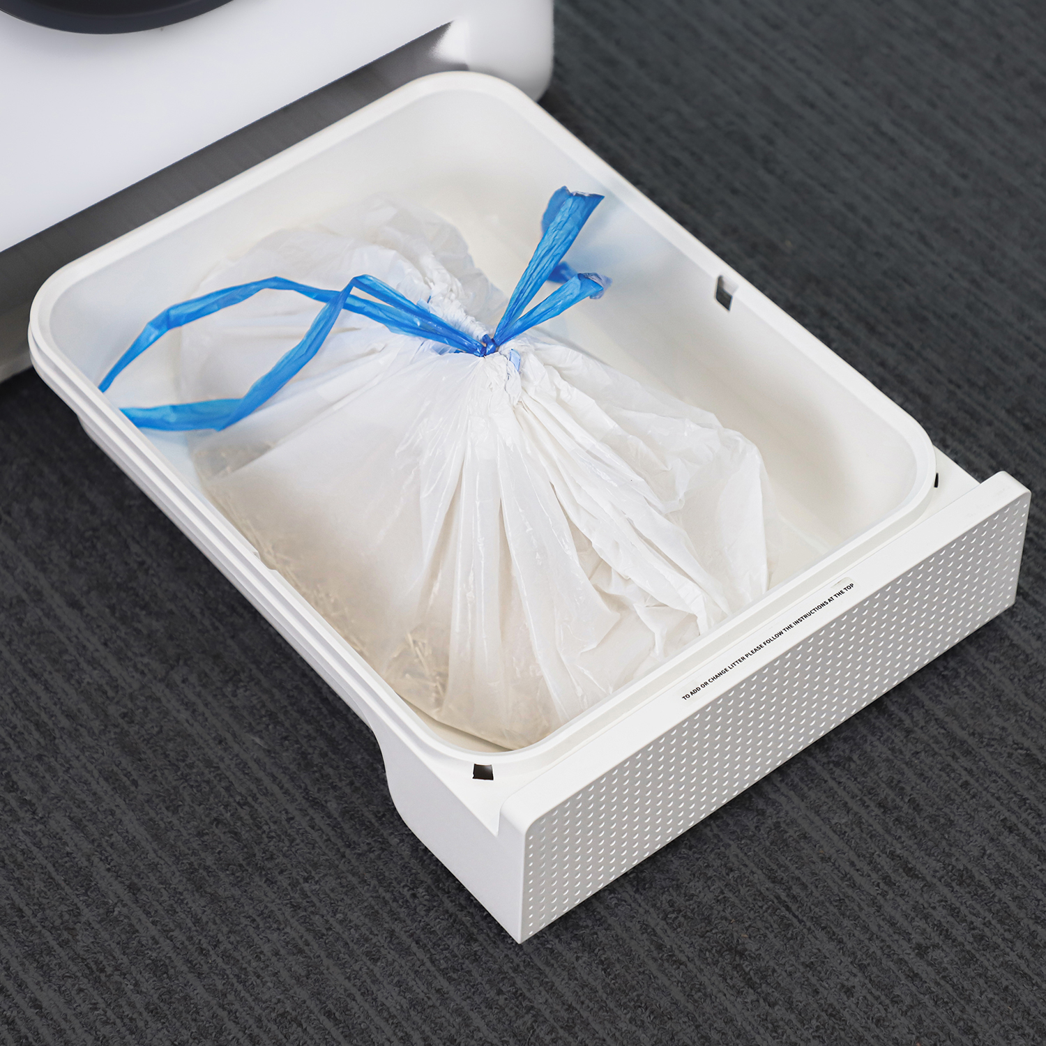 Bidone bianco con sacchetto di plastica all'interno, legato con filo blu. Posizionato su un pavimento grigio.