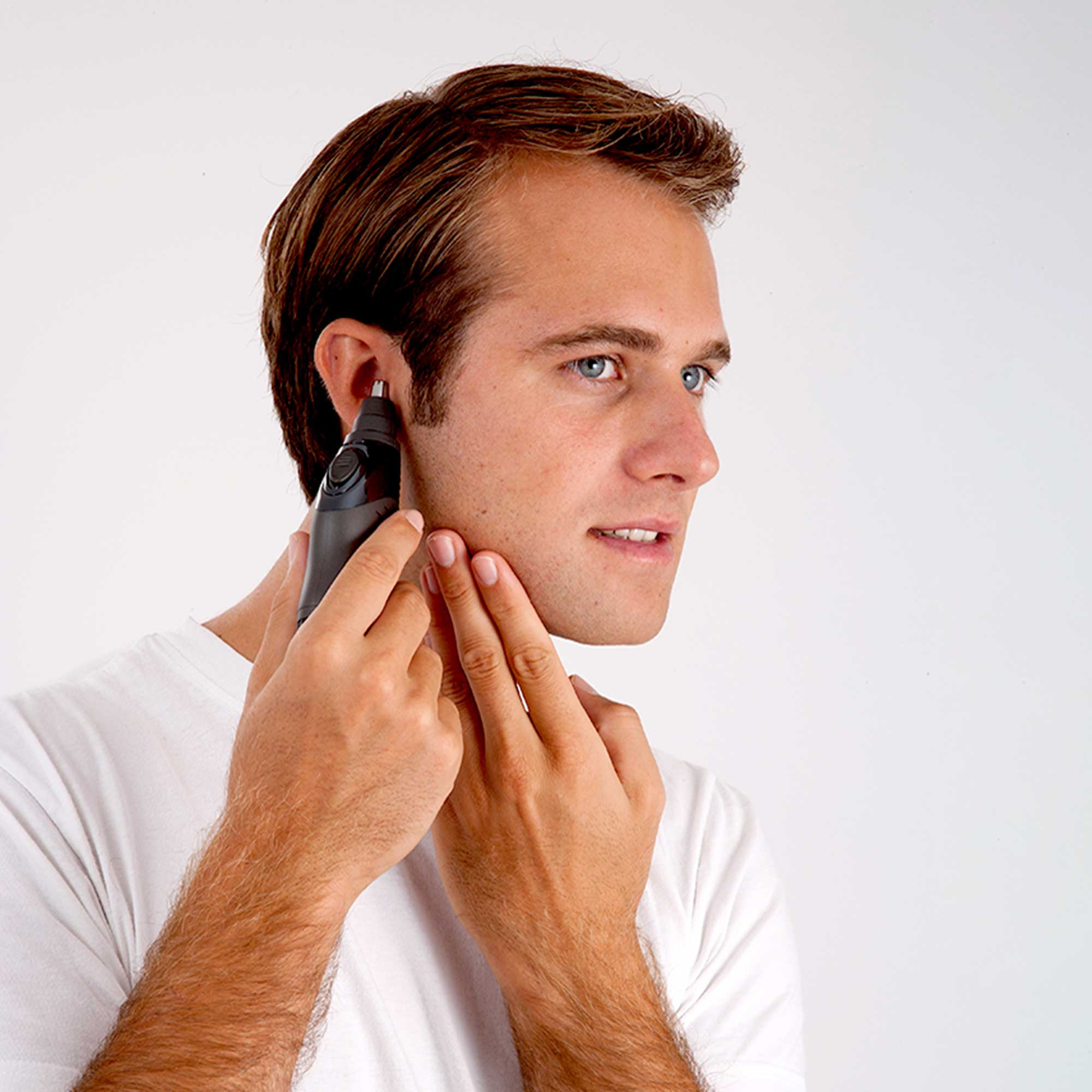 Man trims his ear hair with a black trimmer, side view on a white background.