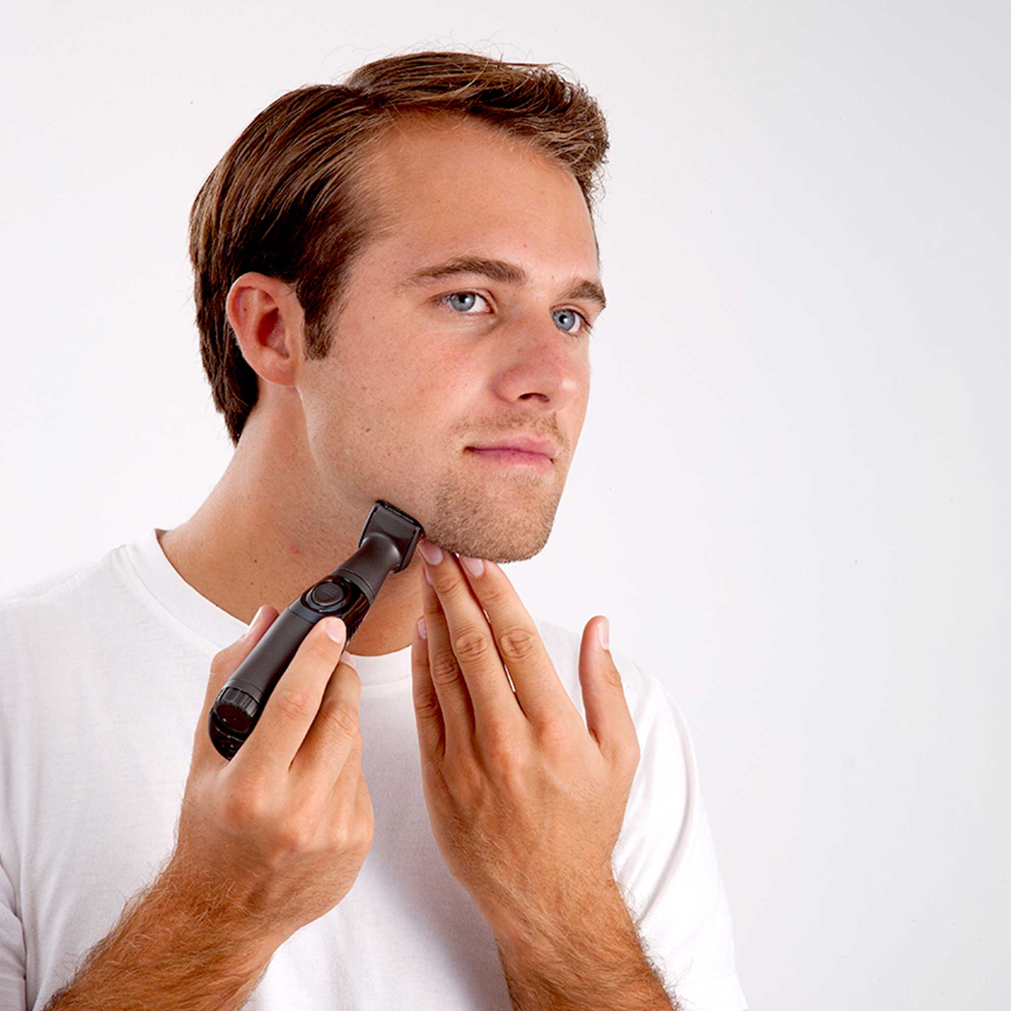 Man using a black beard trimmer on his jawline. He's wearing a white t-shirt. Bright background.