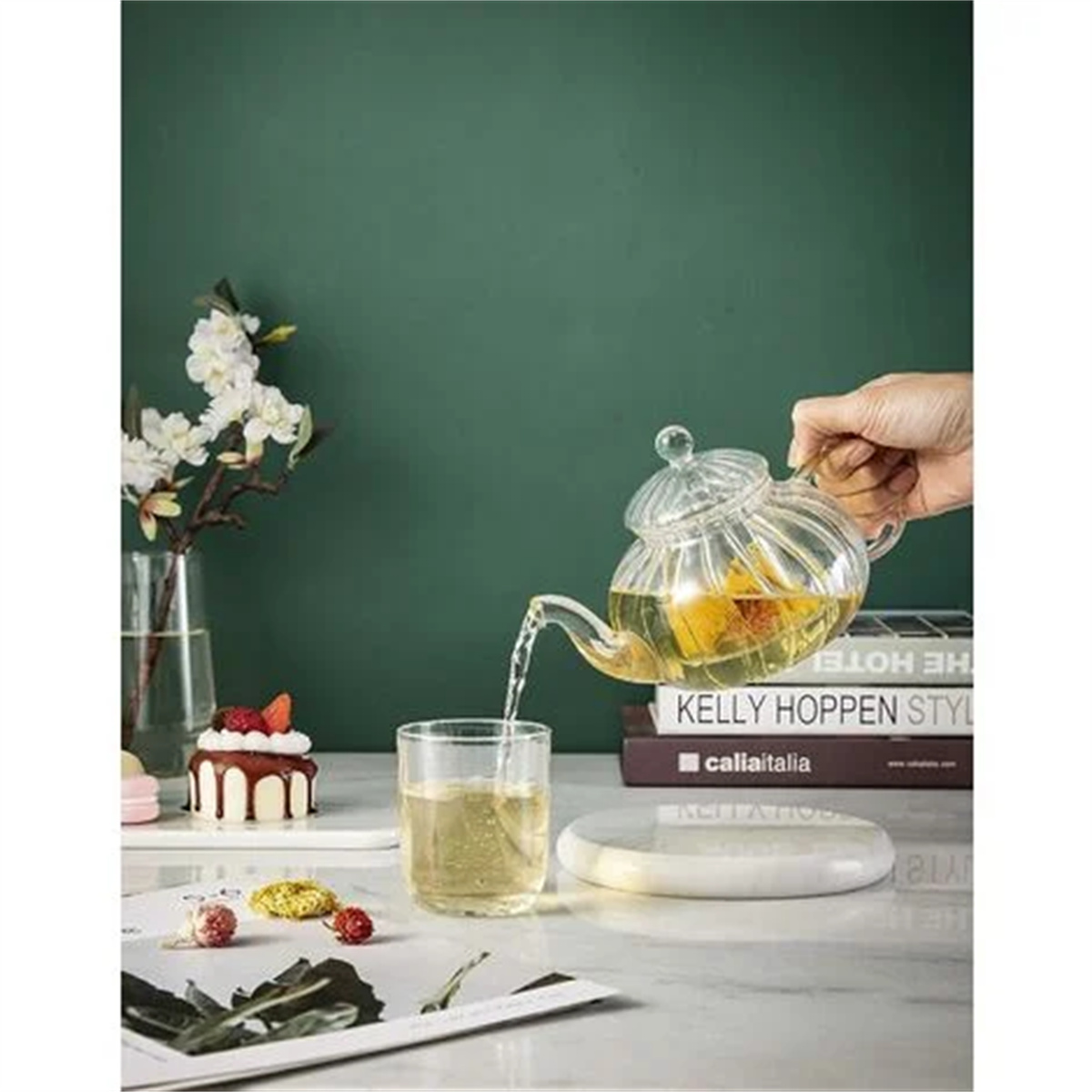 Hand gießt Tee in ein Glas. Teekanne, Bücher und Desserts auf dem Tisch. Grüner Wandhintergrund.