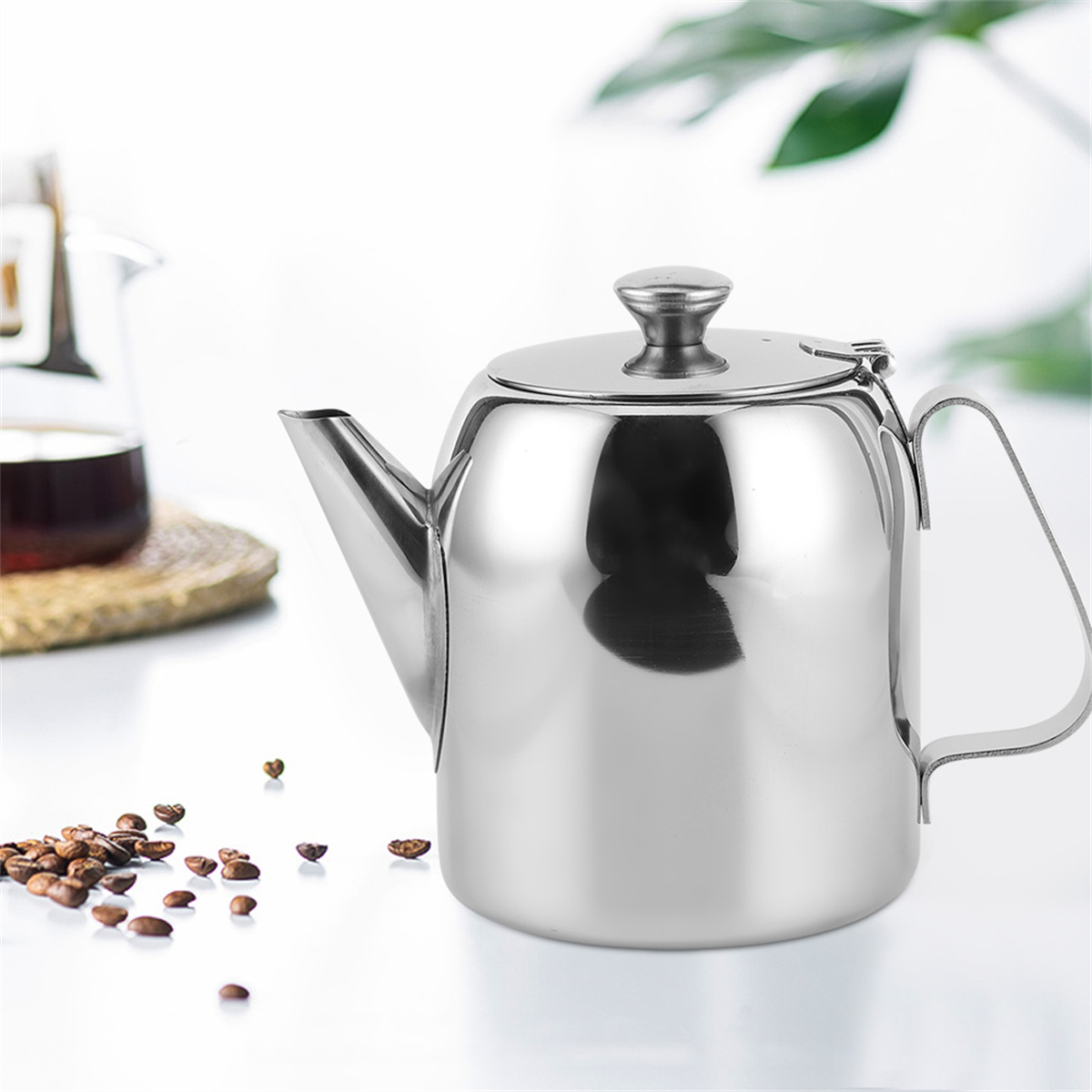 Eine glänzende Teekanne aus Edelstahl mit Deckel und Griff. Kaffeebohnen sind in der Nähe verstreut.