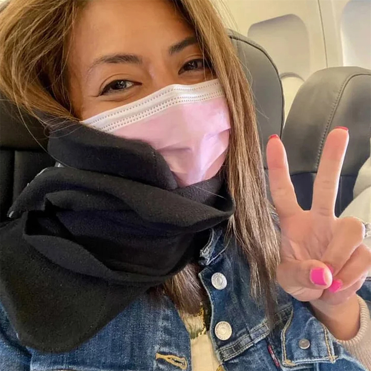 Frau im Flugzeug mit Gesichtsmaske und Schal, macht Friedenszeichen mit pinken Nägeln.