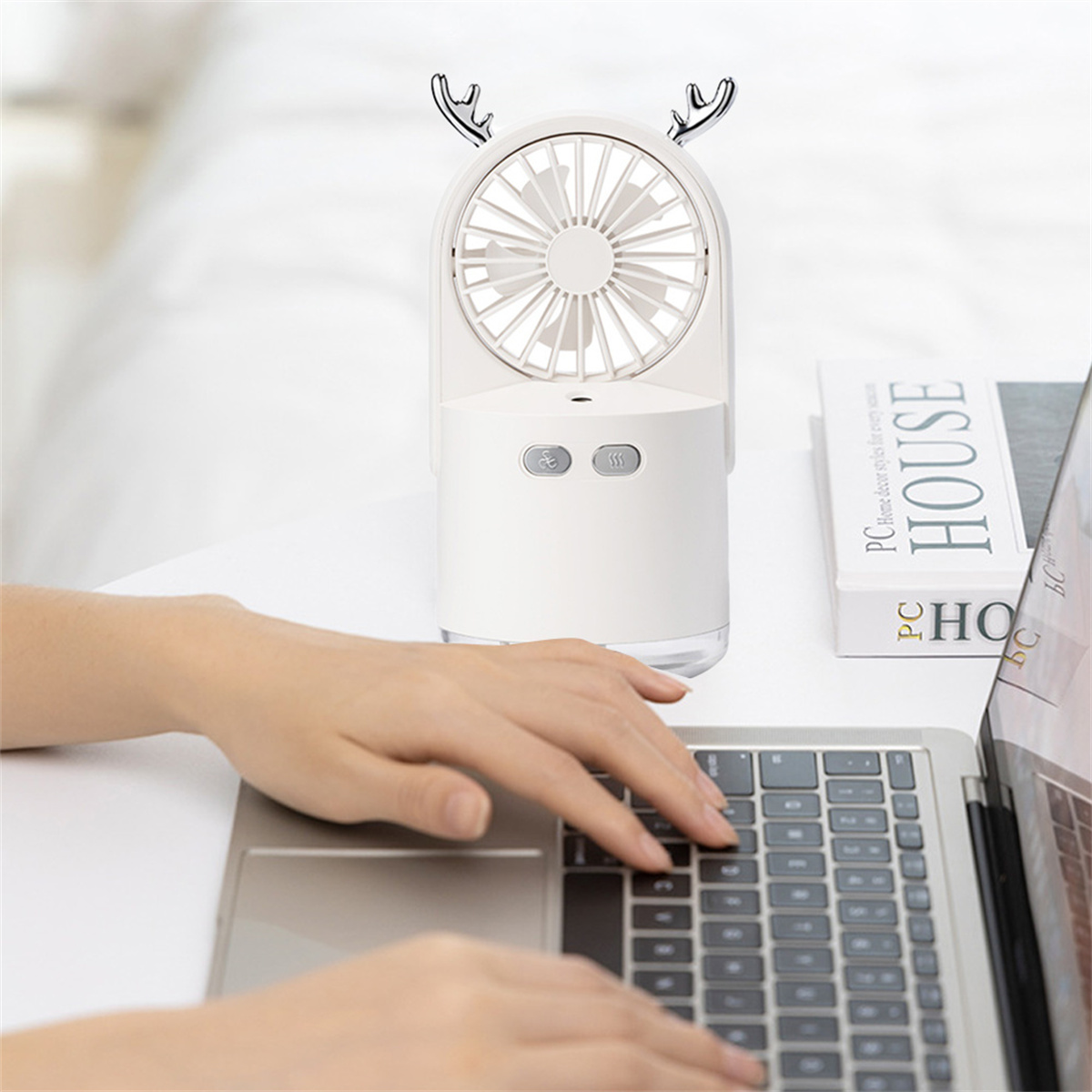 Weißer tragbarer Ventilator mit Hirschgeweih, auf einem Tisch mit Laptop und Büchern.