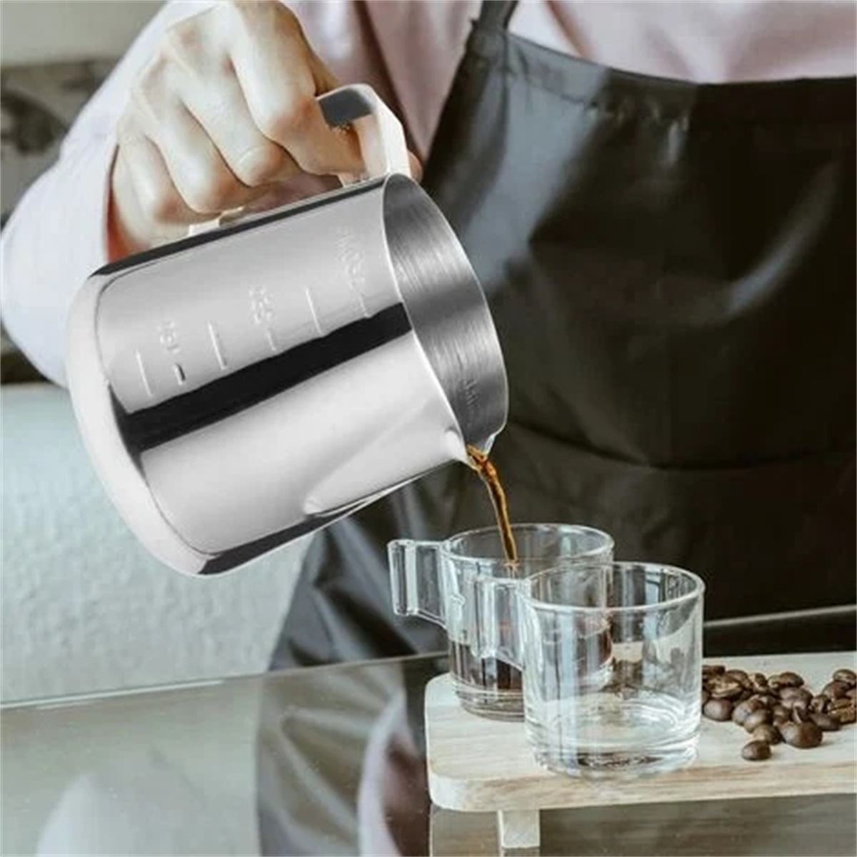 Eine Person gießt Kaffee aus einem Metallkrug in zwei klare Glasbecher auf einem Holzbrett.