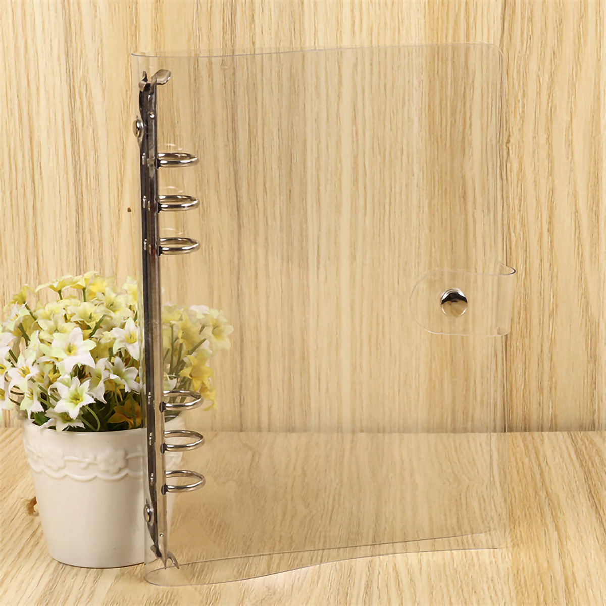 Ein durchsichtiger Ordner mit Metallrücken. Eine Vase mit Blumen steht auf einem Holztisch.