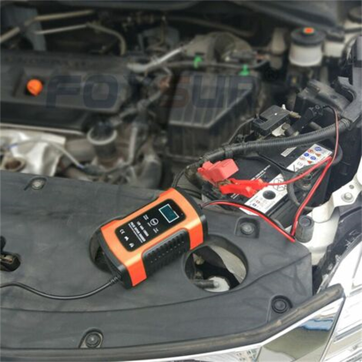 Ein Motorraum mit Batterie und einem Ladegerät mit orangefarbenem und schwarzem Gehäuse, mit roten und schwarzen Klemmen.