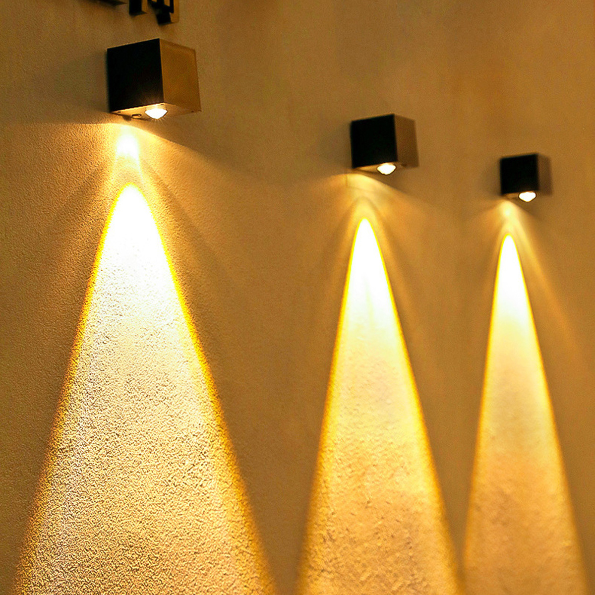 Drei Wandleuchten, die nach unten scheinen. Die Lampen sind schwarz quadratisch und an einer beigefarbenen Wand.