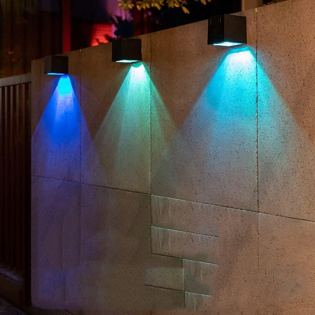 Drei quadratische Außenleuchten an einer Wand, jede mit einem andersfarbigen Lichtstrahl: blau, grün und blau.