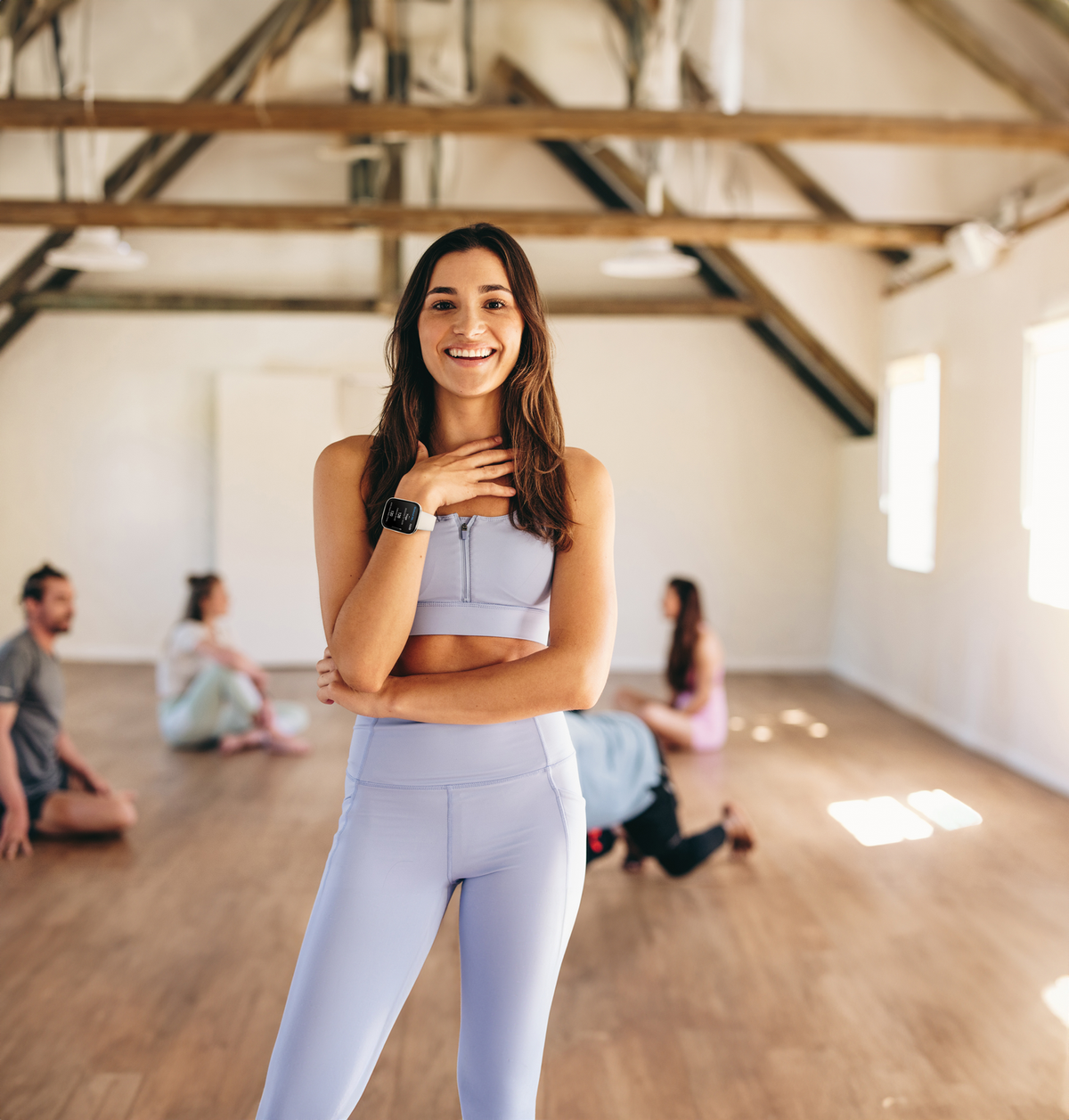 Frau in einem Yoga-Studio, lächelt. Andere Personen im Hintergrund machen Yoga. Holzfußboden, weiße Wände.