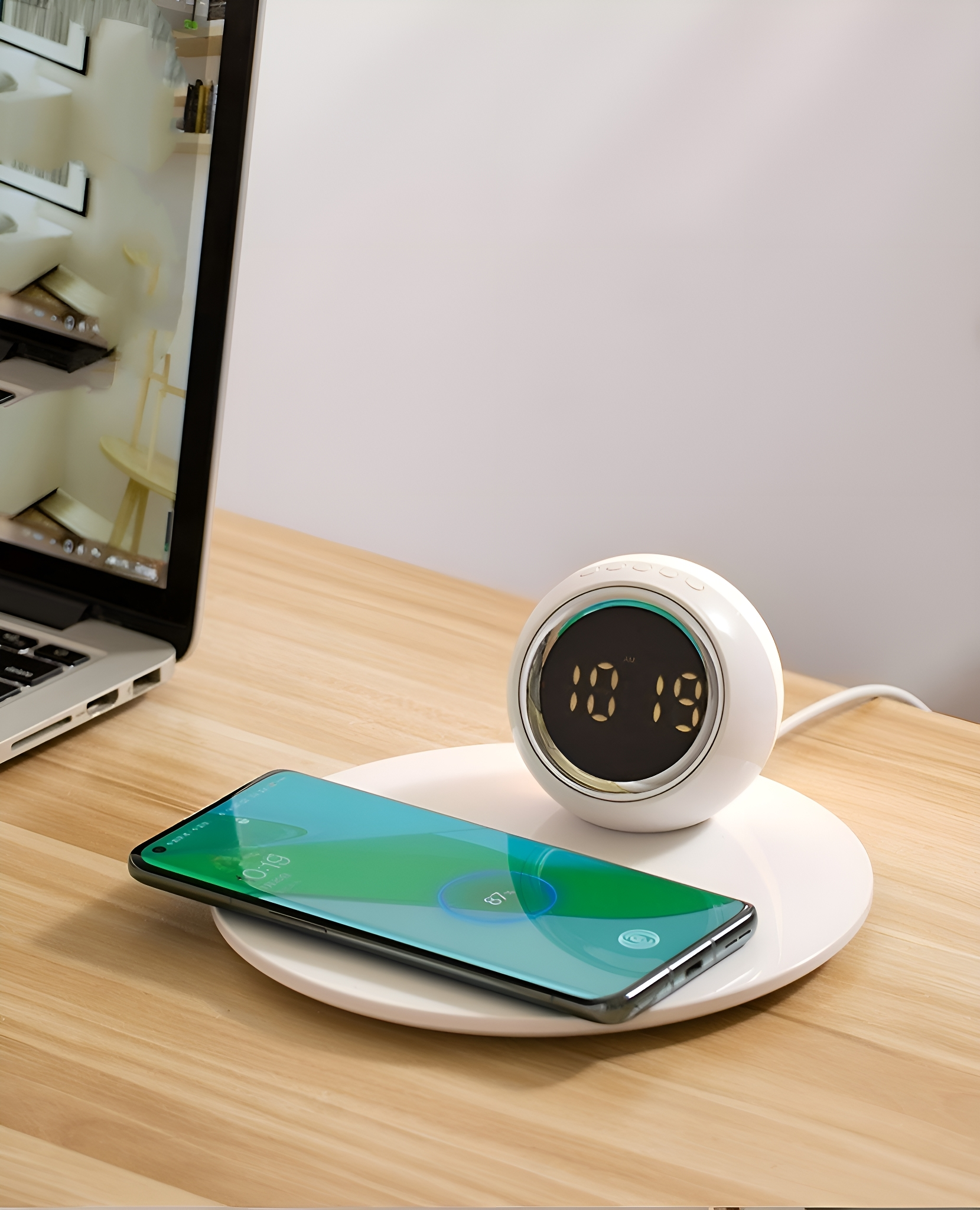 Eine weiße Digitaluhr, ein Telefon wird auf einem weißen Pad geladen. Ein Laptop ist links.