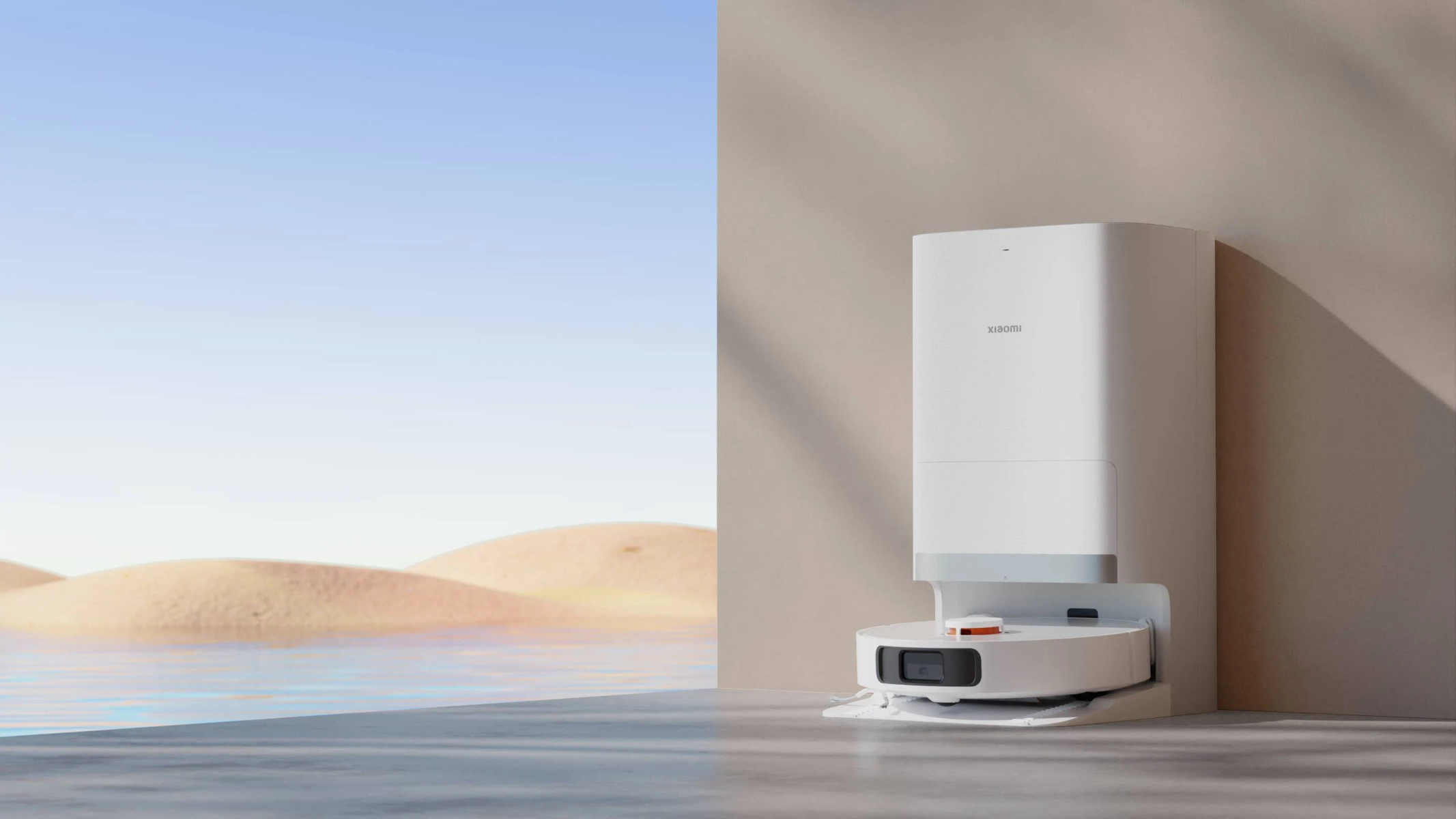 Biały robot odkurzający ze stacją ładującą po prawej stronie, błękitne niebo i wydmy w tle.