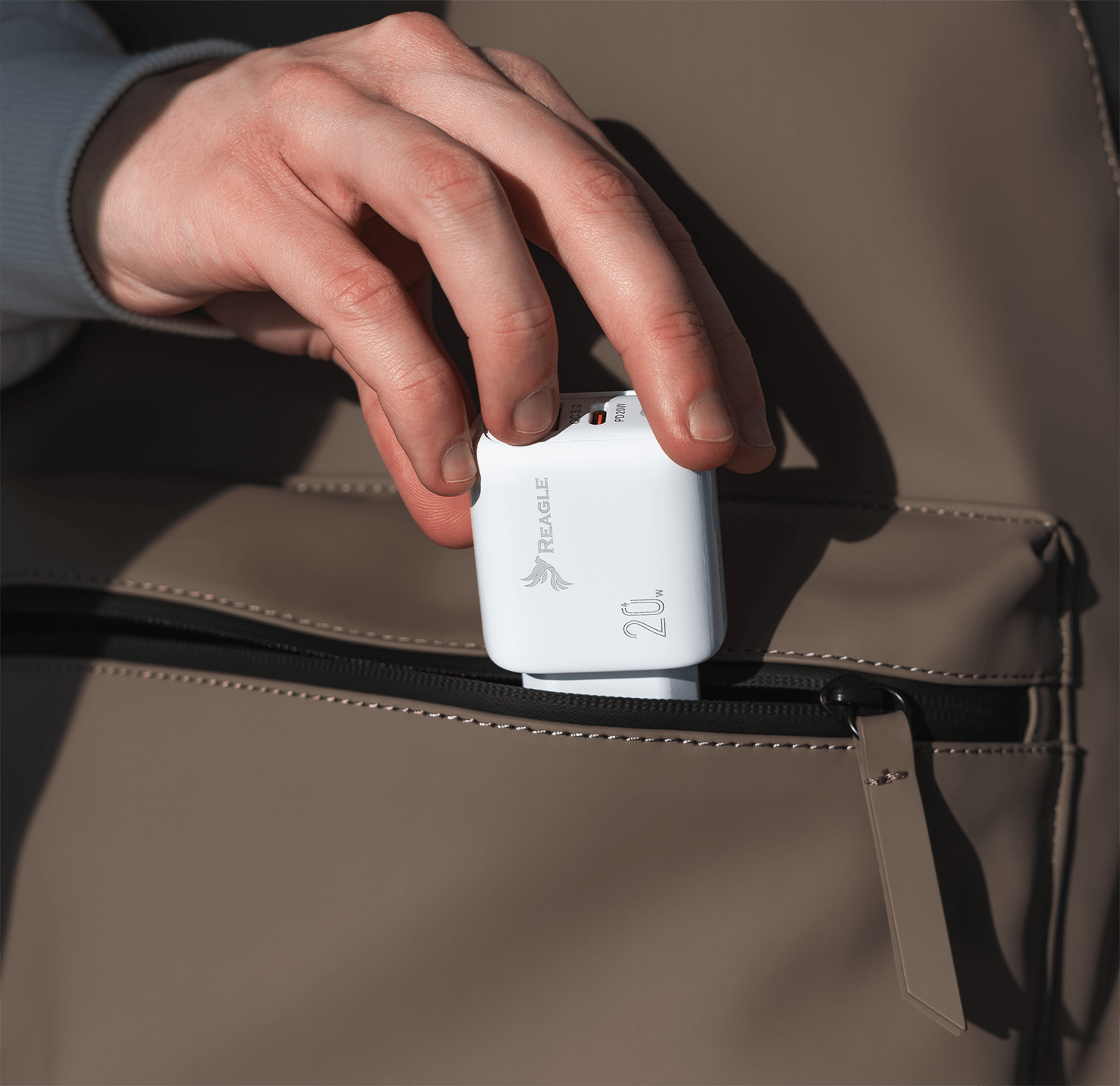 Ręka sięgająca do kieszeni po białą ładowarkę Reagle 20W. Brązowe tło torby.