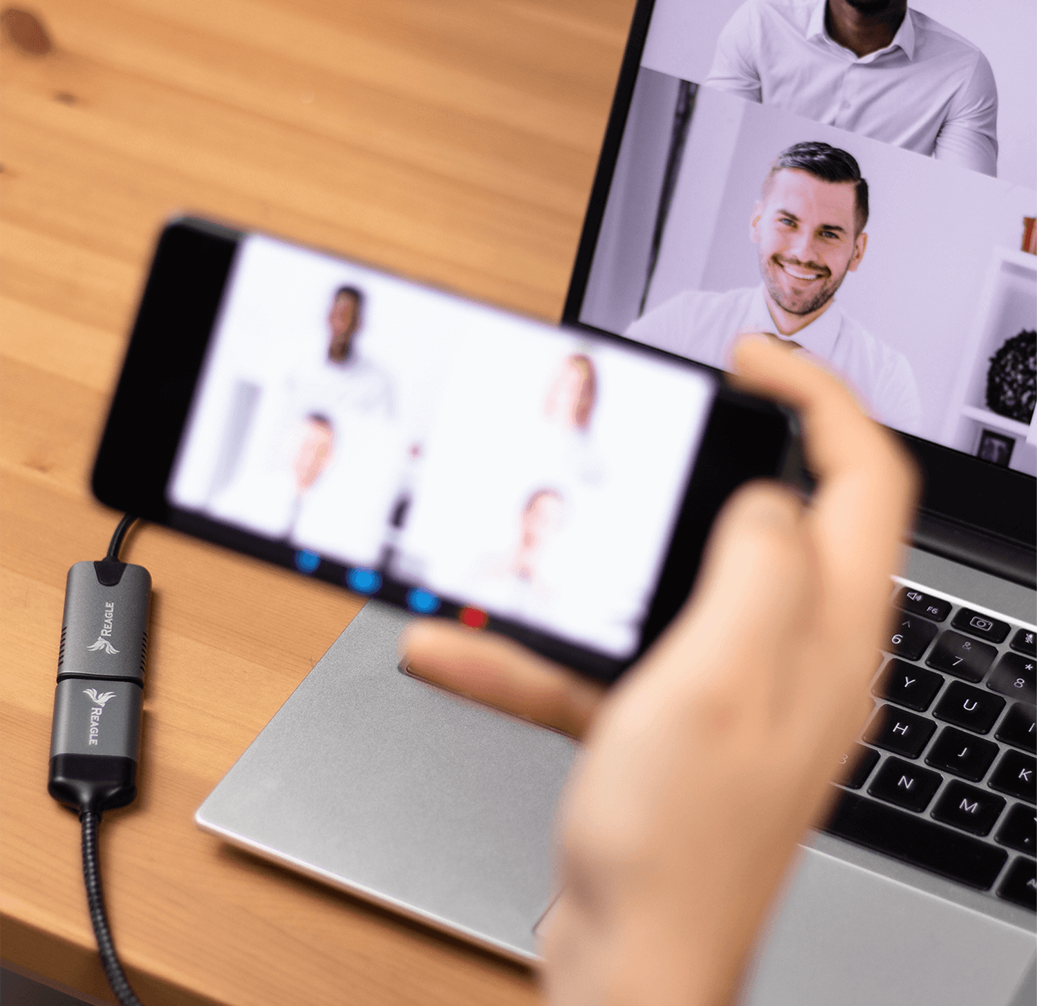 Dłoń trzymająca telefon z rozmową wideo, podłączony do laptopa na drewnianym stole.