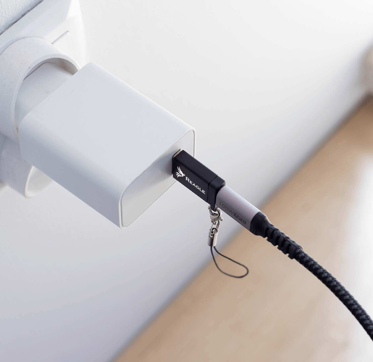 Czarny i srebrny kabel ładujący podłączony do białej ładowarki na jasnej ścianie. Ładowarka ma napis REAGLE.