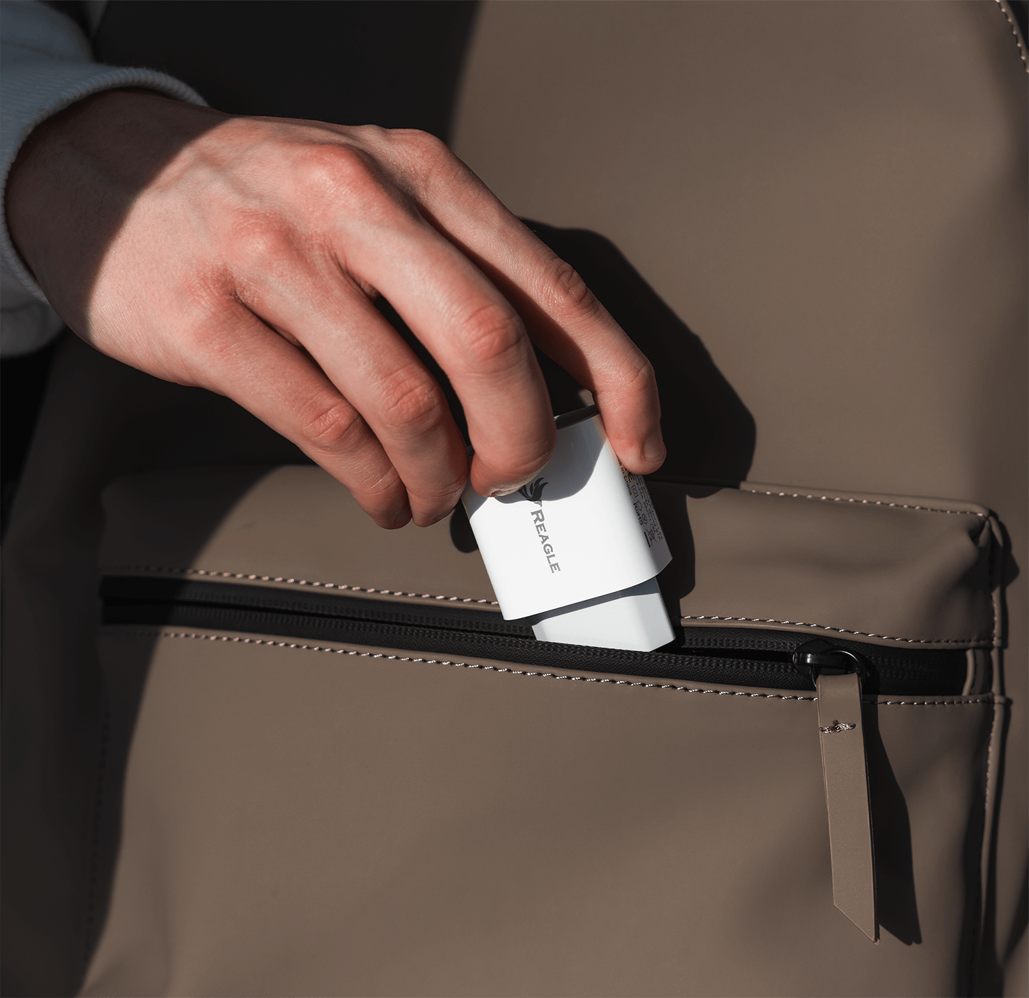 Ręka trzymająca białą ładowarkę Reagle i wkładająca ją do kieszeni na brązowym plecaku.