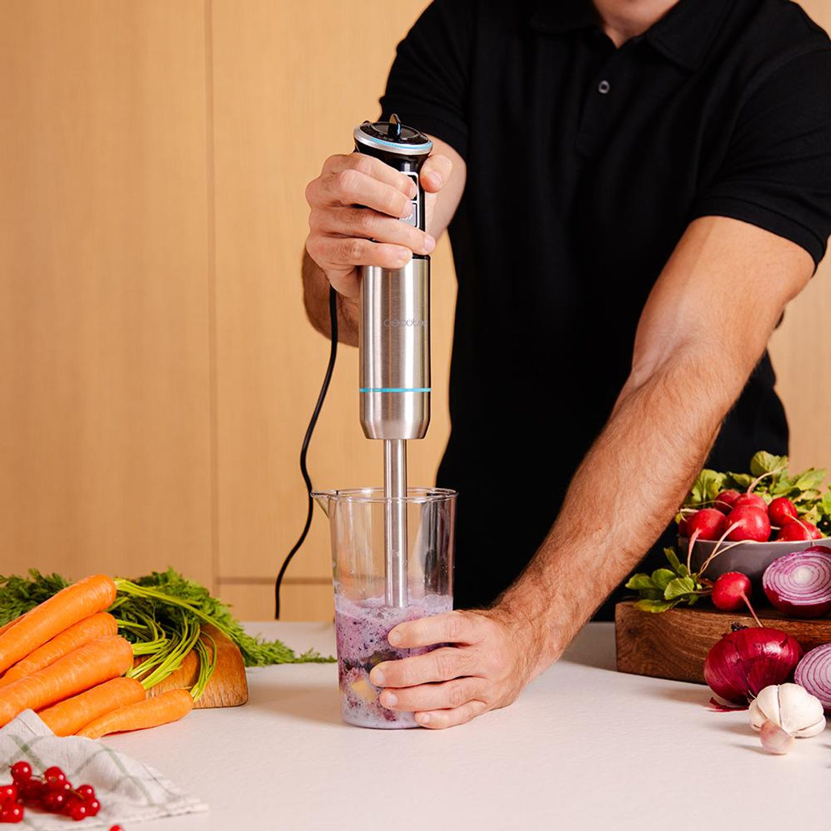 Una persona frulla ingredienti in un bicchiere con un frullatore a immersione. Carote e verdure nella scena.