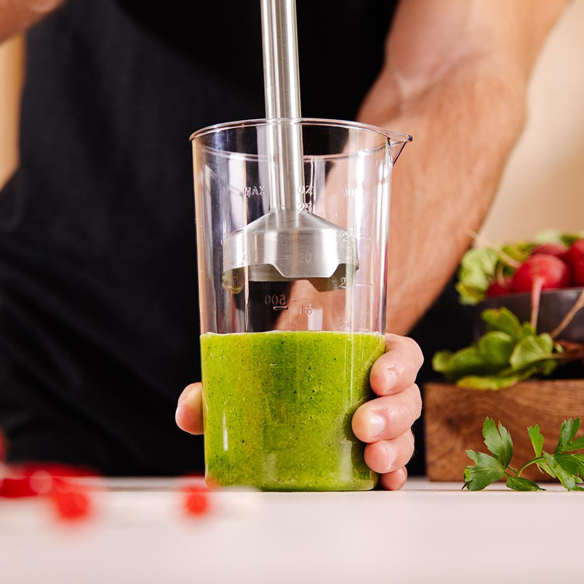 Una mano che tiene un bicchiere trasparente con liquido verde, un frullatore a immersione argentato. Sfondo sfocato.