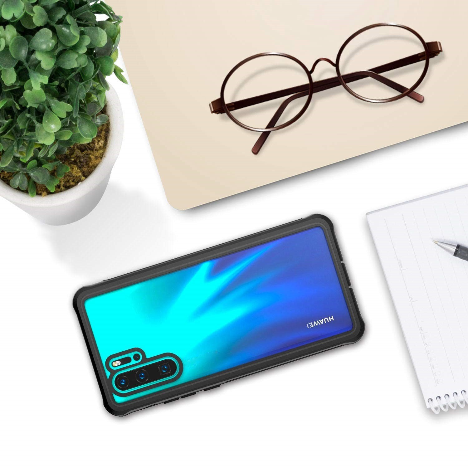 Etui na telefon, okulary, roślina i notatnik na białej powierzchni. Etui czarne z niebieskim gradientem. Brązowe okulary.