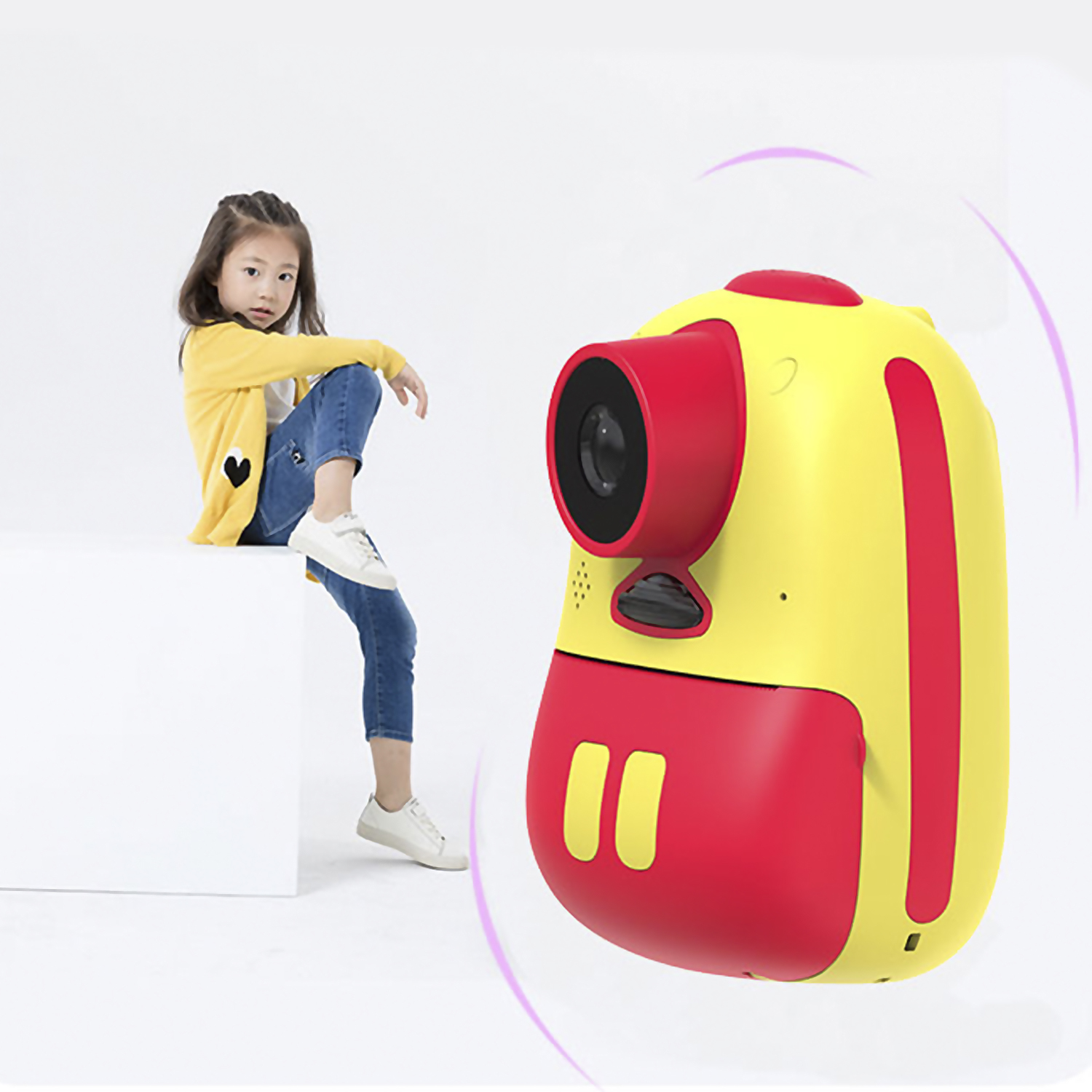 Un niño con chaqueta amarilla y vaqueros azules sentado junto a un juguete con forma de cámara amarillo y rojo.