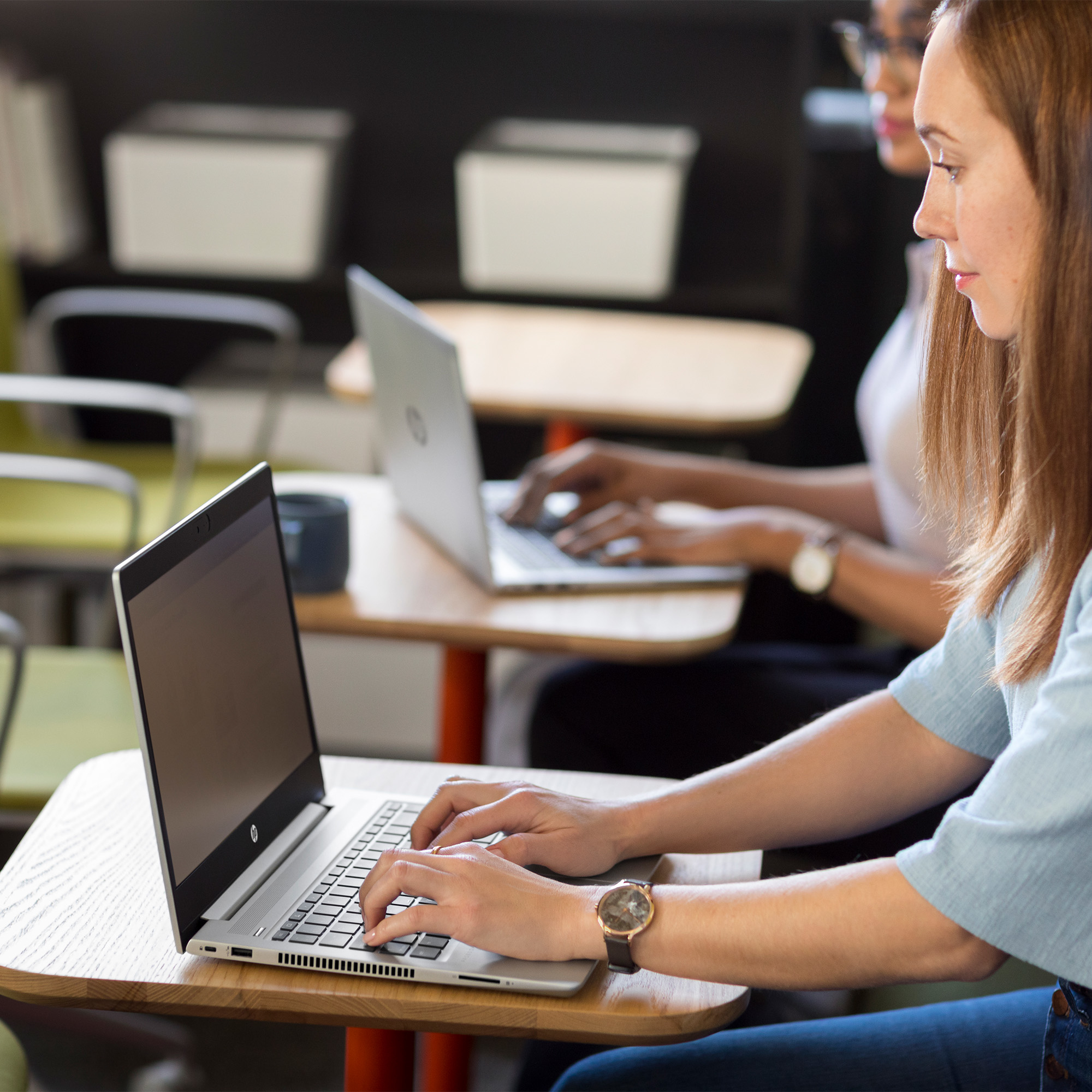 Due donne che lavorano su computer portatili a un tavolo di legno in un ambiente ufficio moderno.
