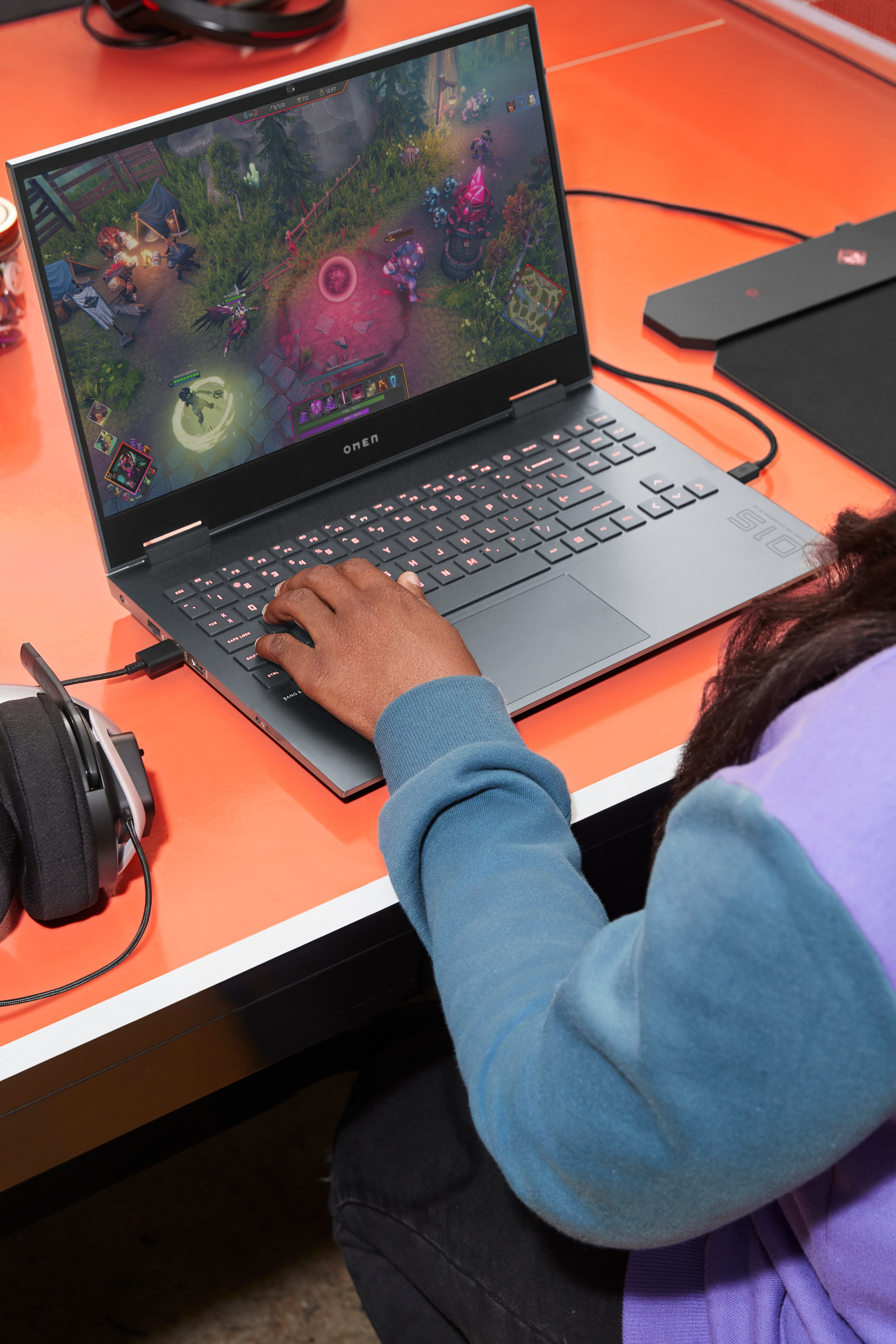 Osoba korzystająca z laptopa z grą wyświetlaną na ekranie, obok zestawu słuchawkowego.