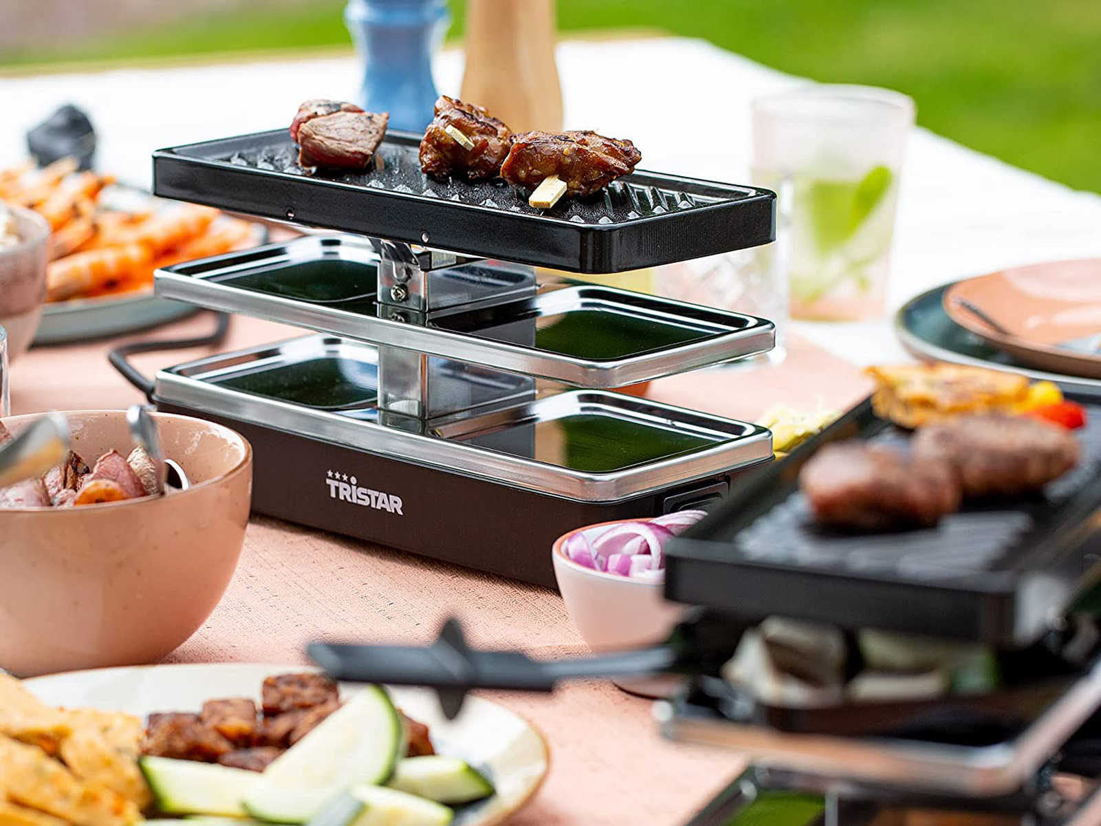 Griglia per raclette con carne e verdure su un tavolo, con altri cibi e bevande sullo sfondo.