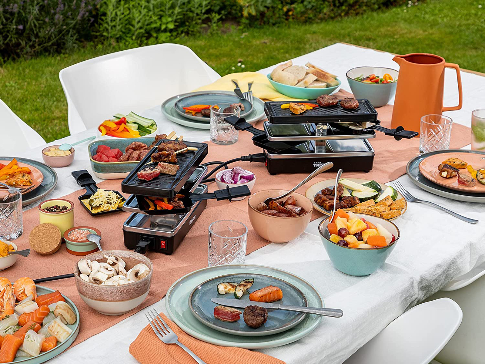 Tavolo da pranzo all'aperto con vari cibi e griglie per raclette. I piatti includono carni, verdure e frutta.