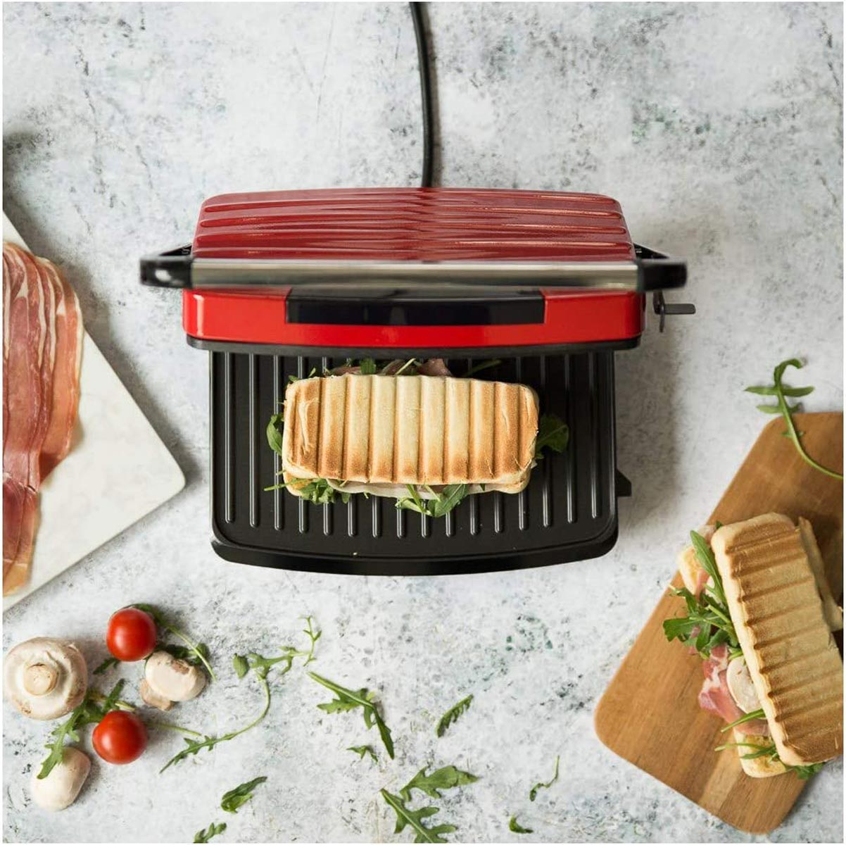 Eine rote Panini-Presse mit einem Sandwich auf dem Grill. Zutaten umgeben die Presse. Es gibt Tomaten und Rucola.