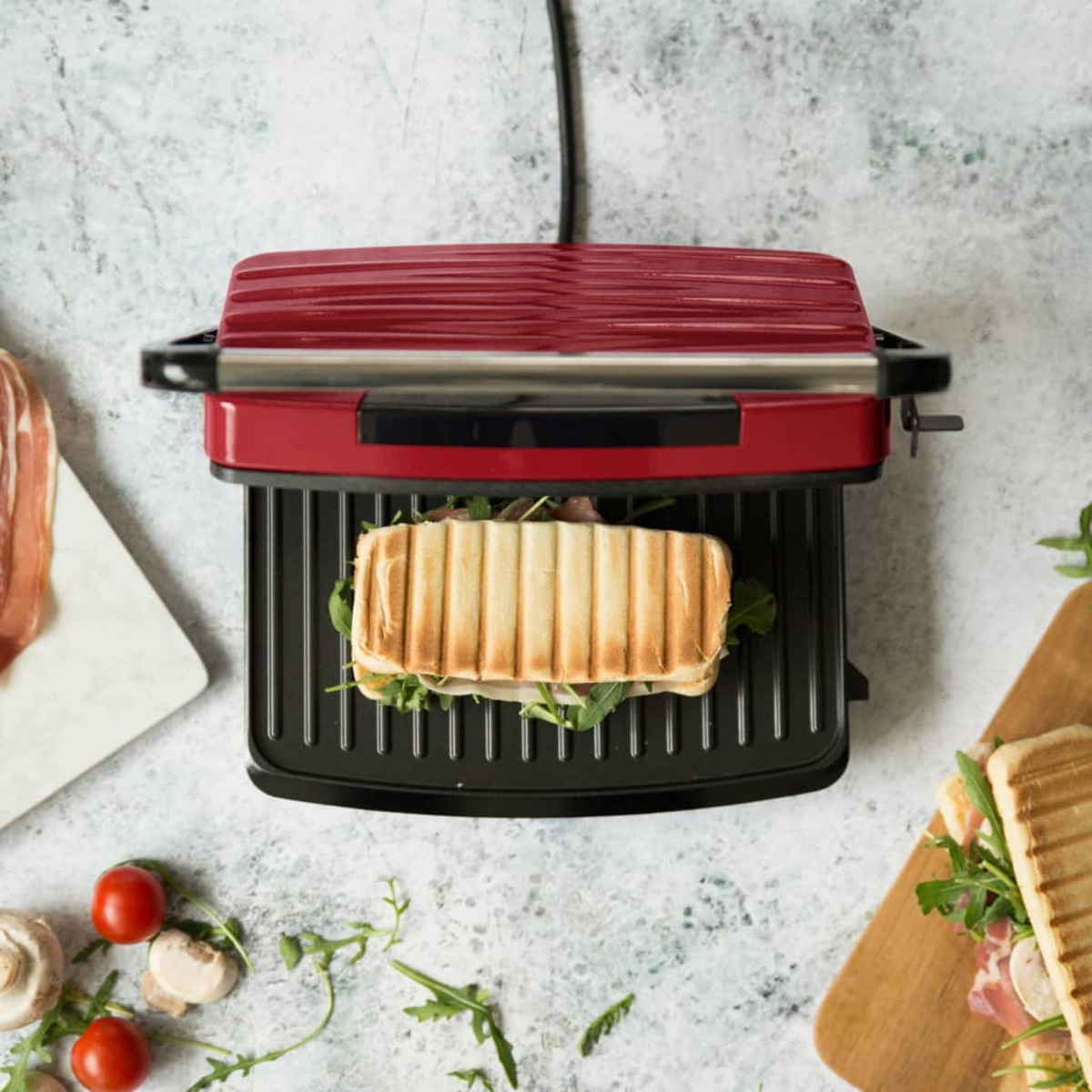 Eine rote Panini-Presse mit einem Sandwich auf dem Grill. Tomaten, Rucola und Sandwich liegen auf der Theke.