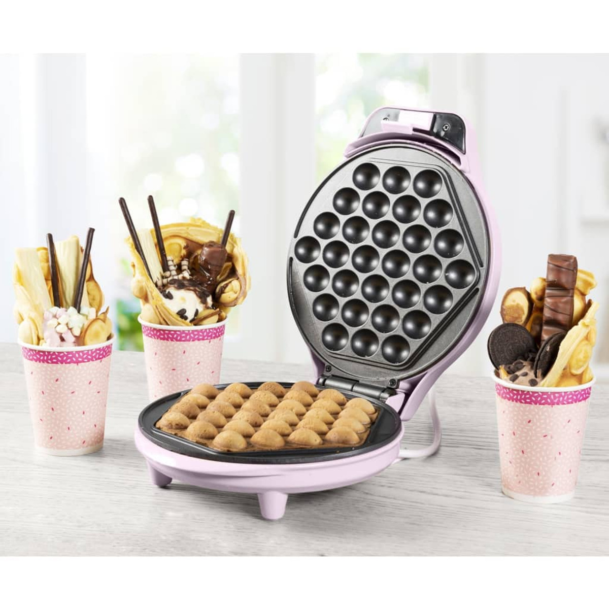 Macchina per waffle rosa con waffle. Diversi coni con condimenti ai lati.