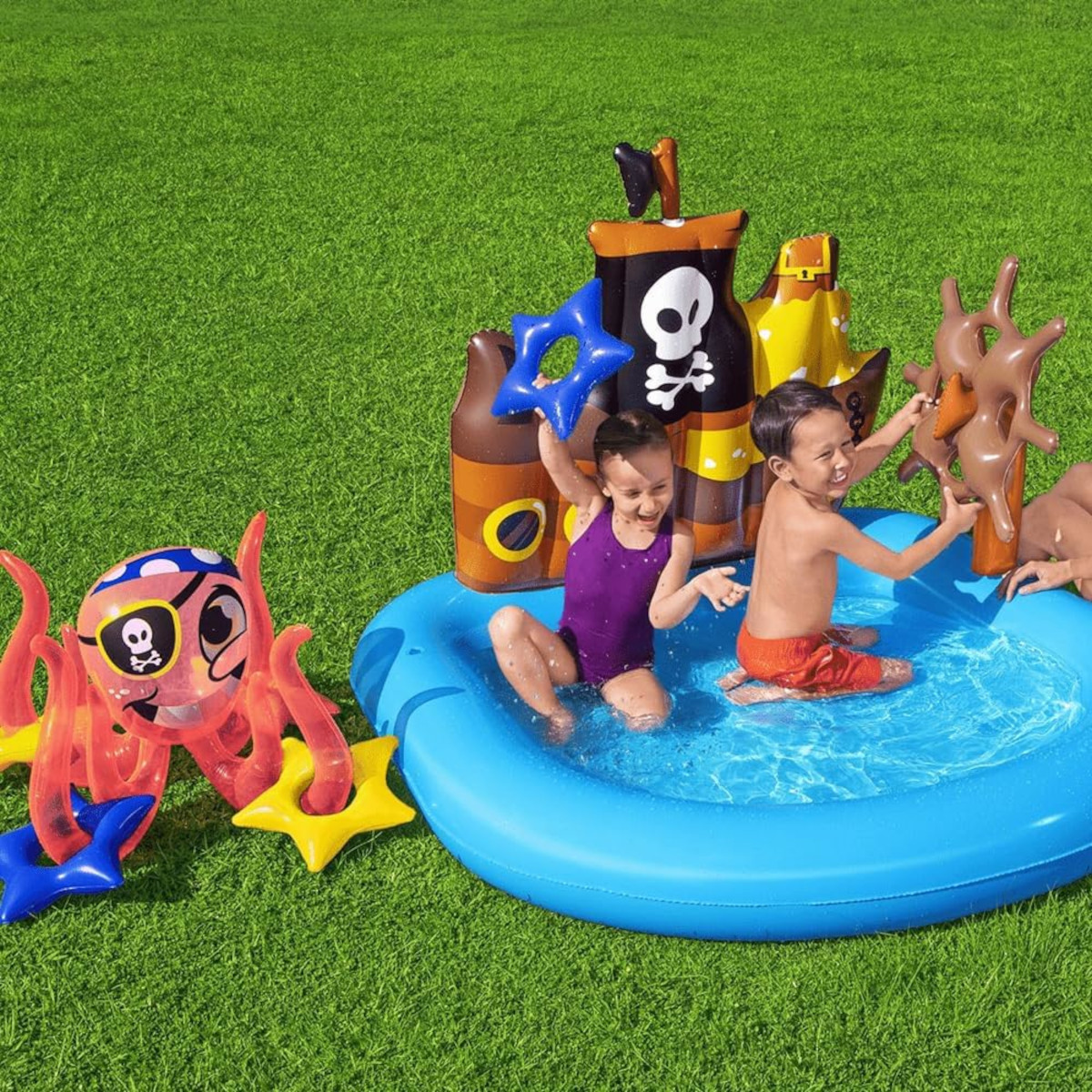 Due bambini giocano in una piscina gonfiabile blu con decorazioni a tema pirata sull'erba verde.