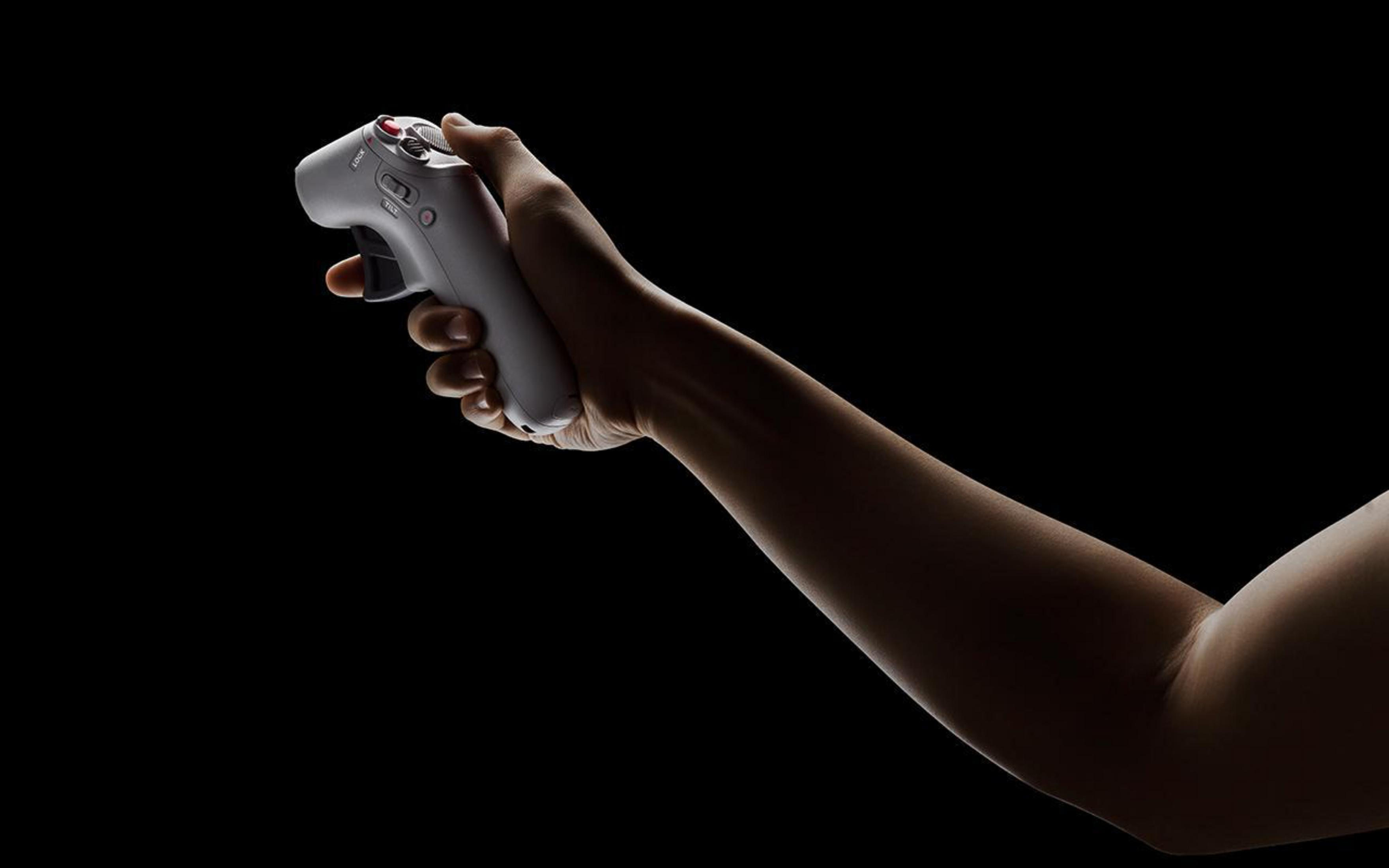 Ręka trzymająca szary kontroler do gier na czarnym tle. Kontroler jest oświetlony z boku.