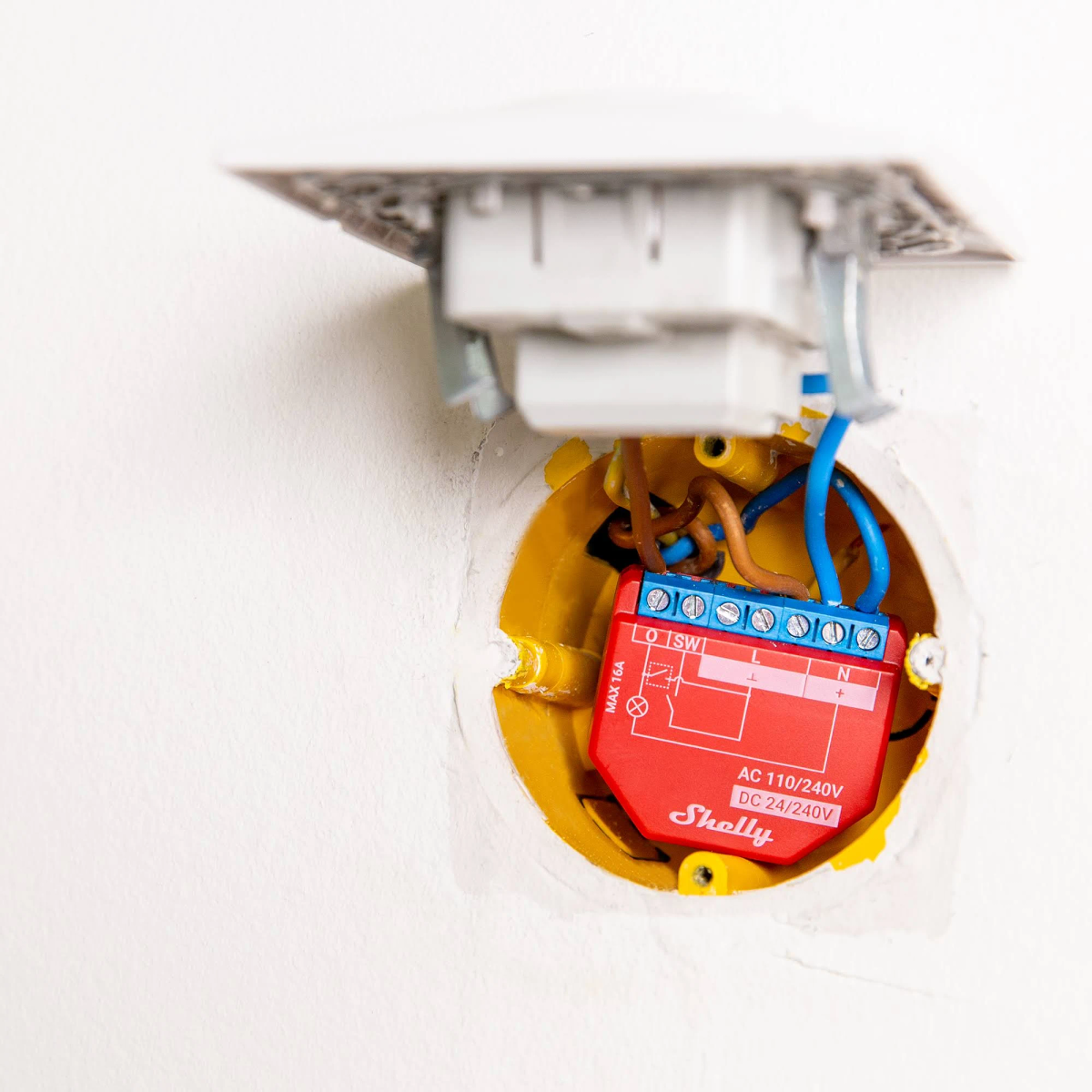 Dispositivo domótico rojo, cableado en una caja eléctrica amarilla, montado en un hueco de pared blanco.
