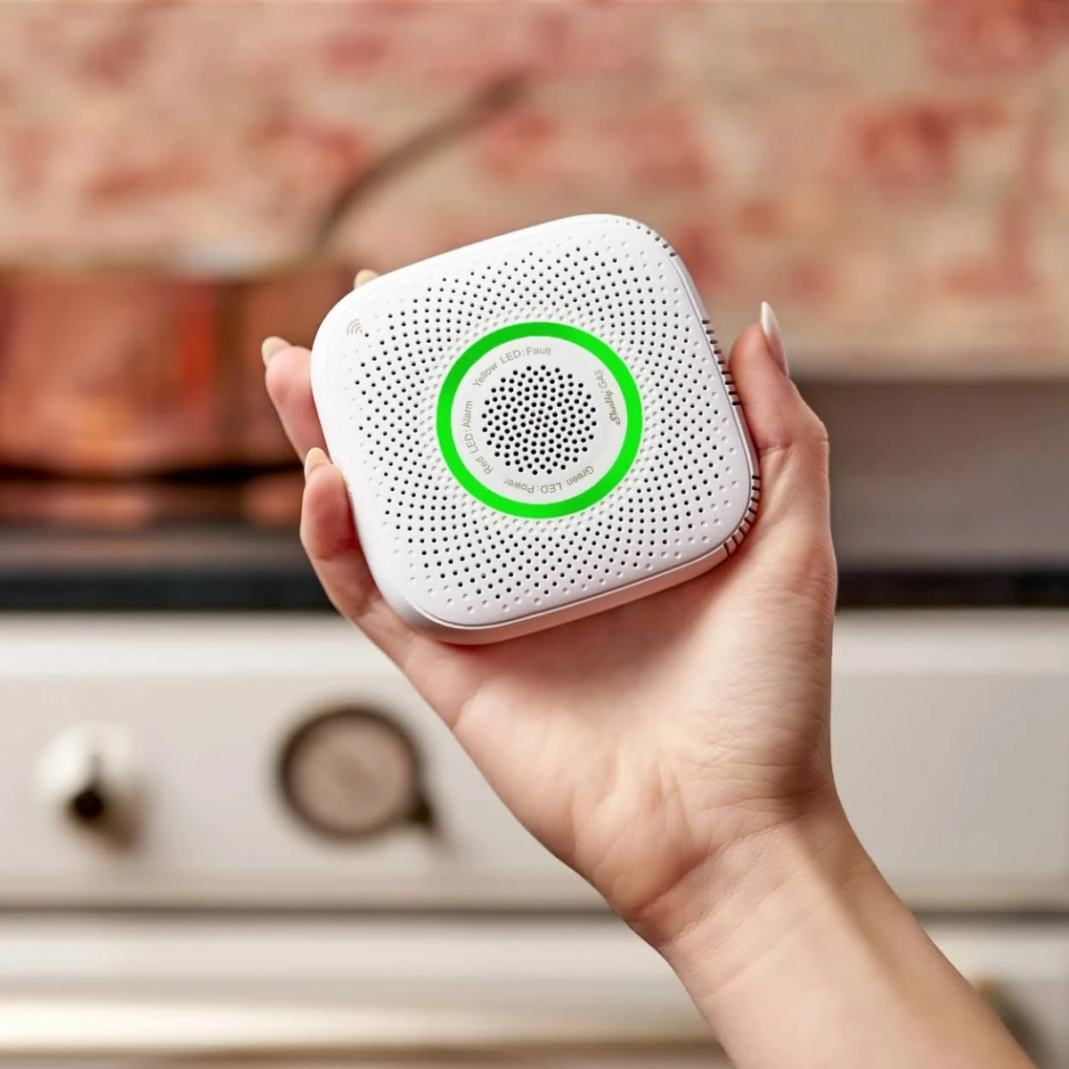 Biały, kwadratowy czujnik dymu. Trzyma go dłoń. Zielony pierścień wokół środka.