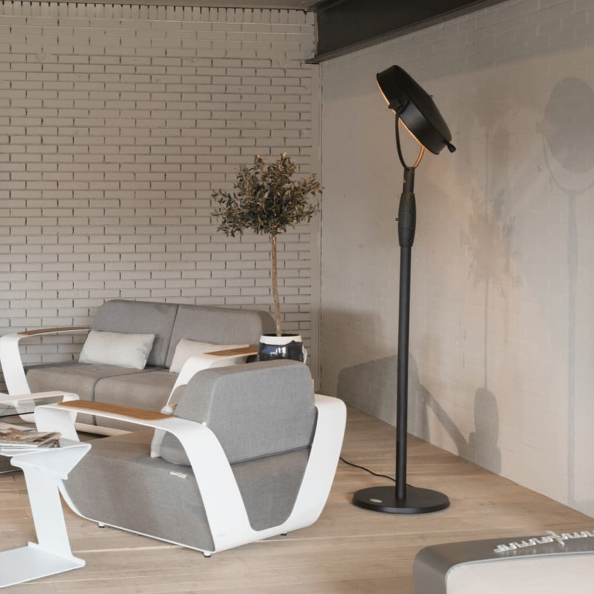 Ein modernes Wohnzimmer mit grauem Sofa, Sessel und schwarzer Stehleuchte. Eine Backsteinmauer im Hintergrund.