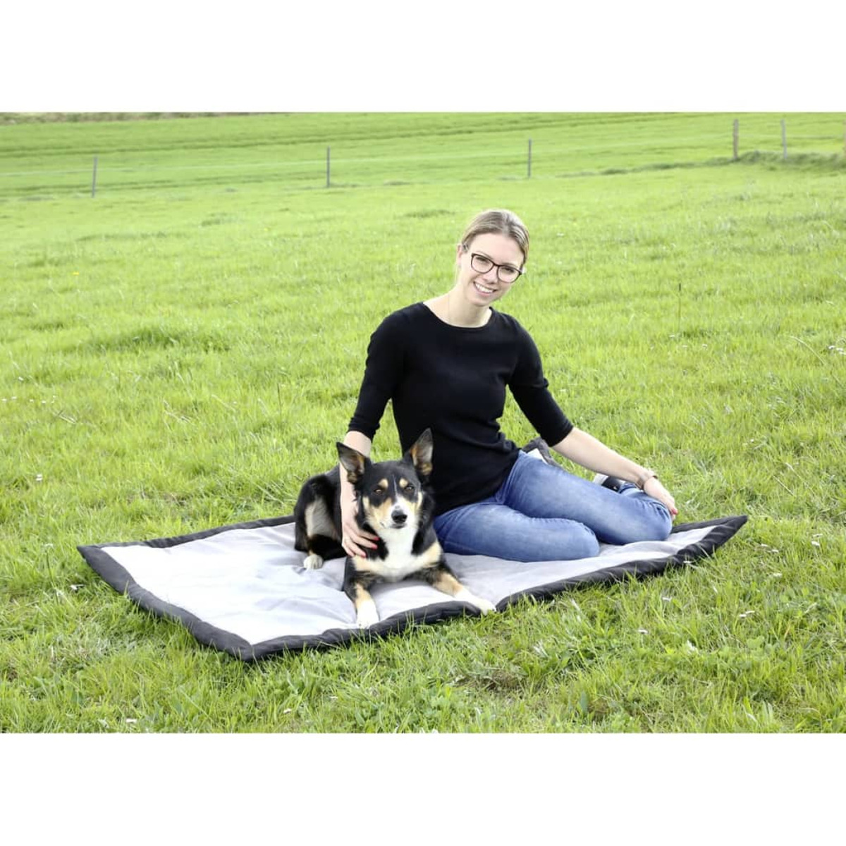 Eine Frau und ein Hund auf einer Decke auf einer Wiese. Die Frau trägt eine Brille und Jeans.