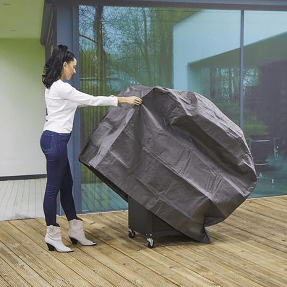 Eine Frau, die einen schwarzen Grill mit einer Plane auf einem Holzdeck abdeckt.