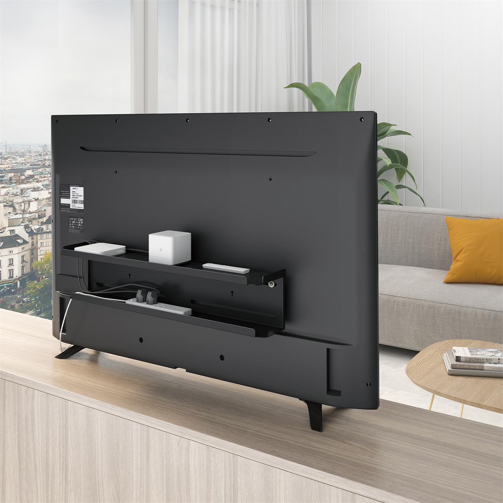Retro di una TV nera con mensola collegata. La mensola contiene un dispositivo bianco e un telecomando. Divano, tavolo e finestra sullo sfondo.