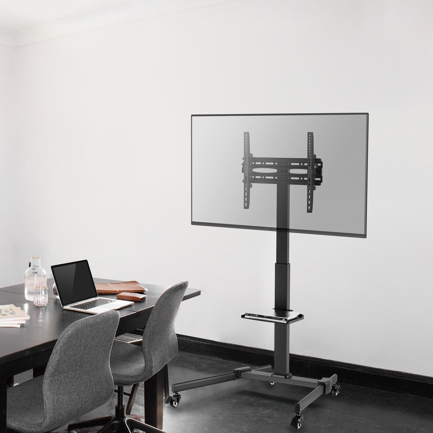 A TV on a stand with wheels is placed in a room. A laptop is on a table next to two grey chairs.
